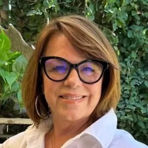 Close-up of a woman with shoulder-length light brown hair, wearing black glasses and a white shirt, smiling outdoors with green foliage in the background.