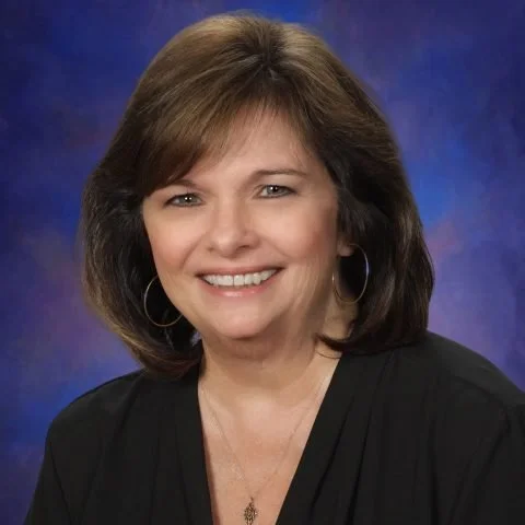 A woman with shoulder-length brown hair, wearing a black top and hoop earrings, smiling against a blue and purple mottled background.