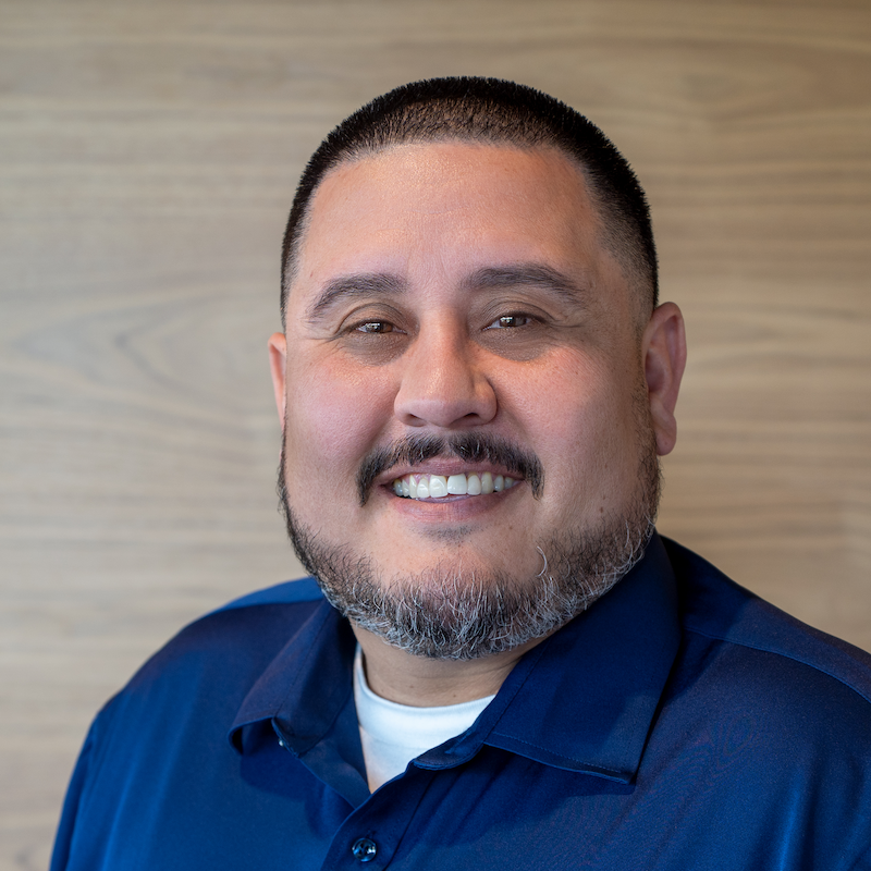 Smiling man with short black hair and beard wearing a navy blue shirt against a light wood background.
