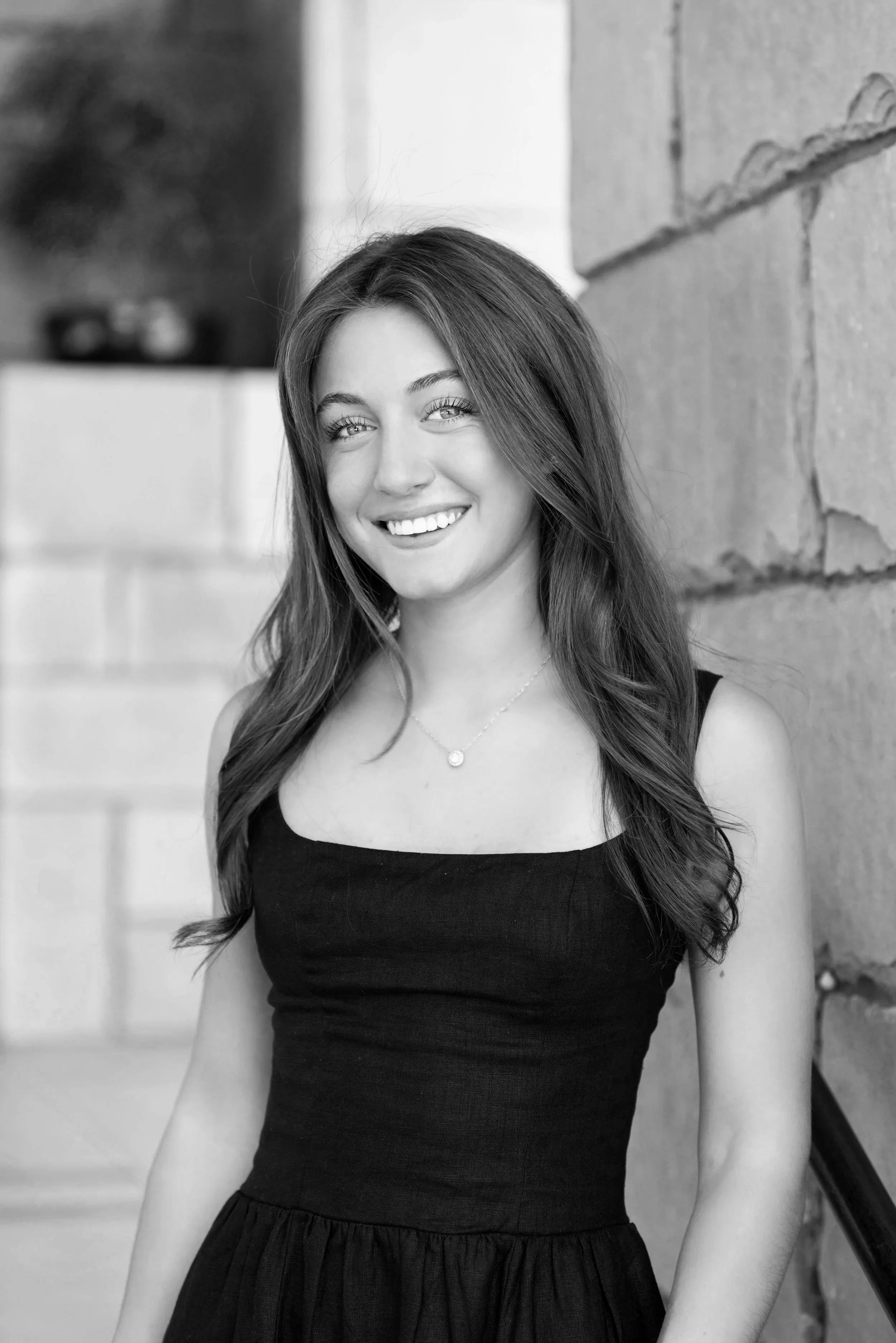Black and white photo of a young woman with long hair smiling, wearing a sleeveless black top and a necklace, standing outdoors against a brick wall.