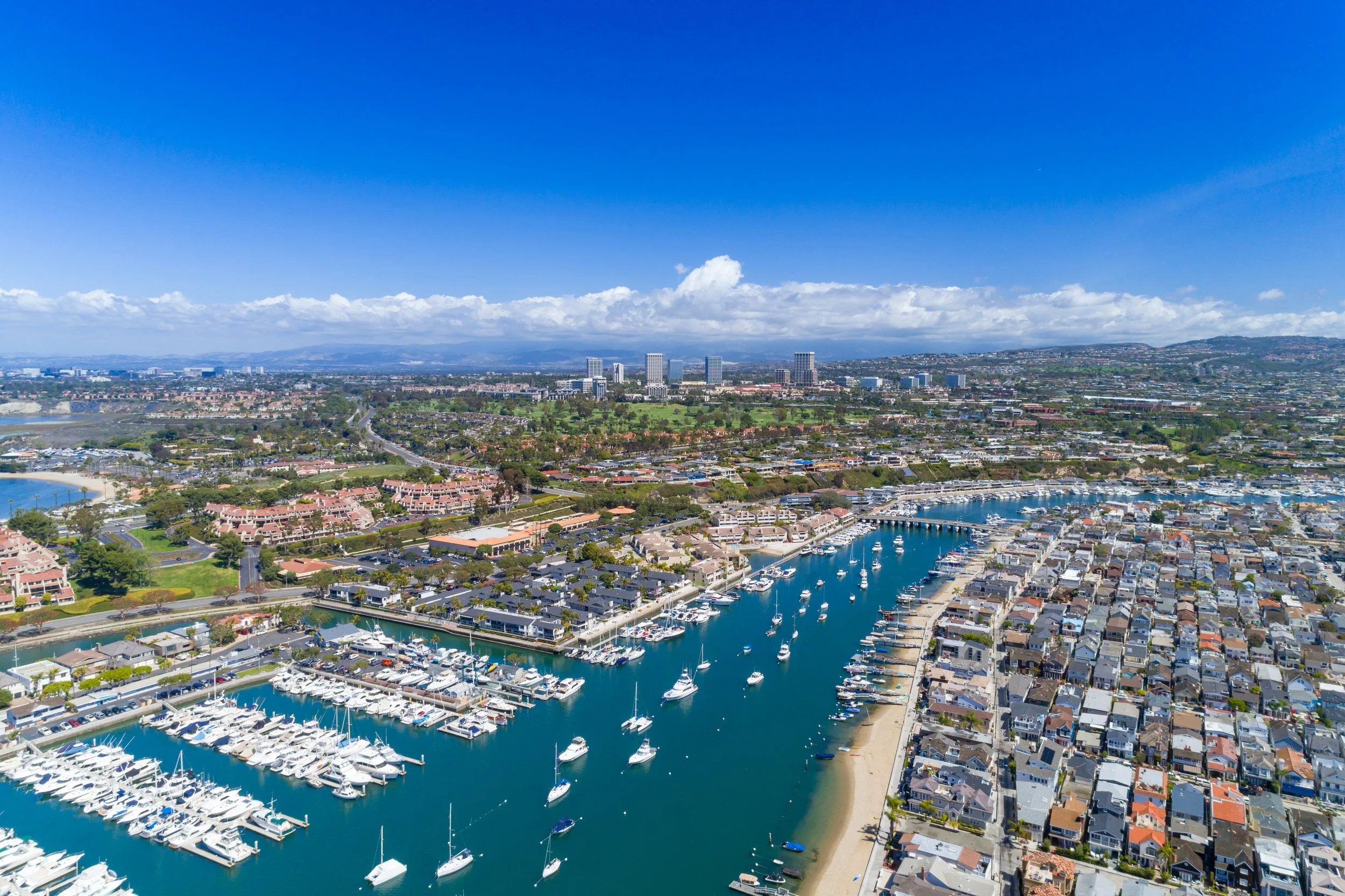Aerial view of a coastal city with a marina filled with boats, residential neighborhoods, and a downtown area with high-rise buildings under a clear blue sky.