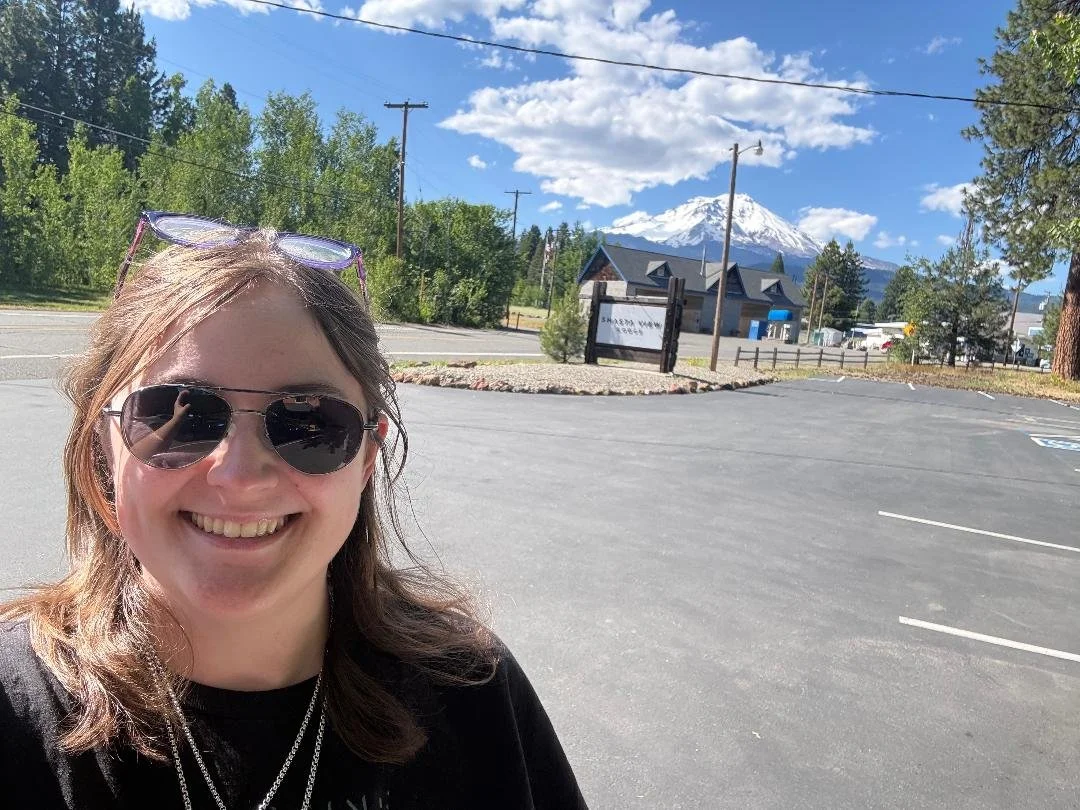 woman smiling wearing sunglasses in an outdoor parking lot with trees, a sign, a house, and Mount St. Helens in the background.
