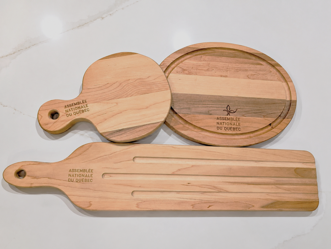 Set of three wooden serving boards, two round and one rectangular, engraved with 'Assemblée Nationale du Québec' and a small plant logo, on a white surface.