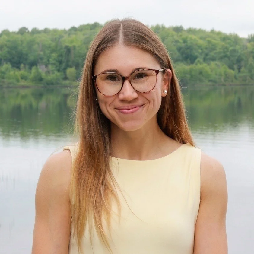 A woman with long brown hair, glasses, and pearl earrings smiling outdoors near a lake, with green trees in the background.