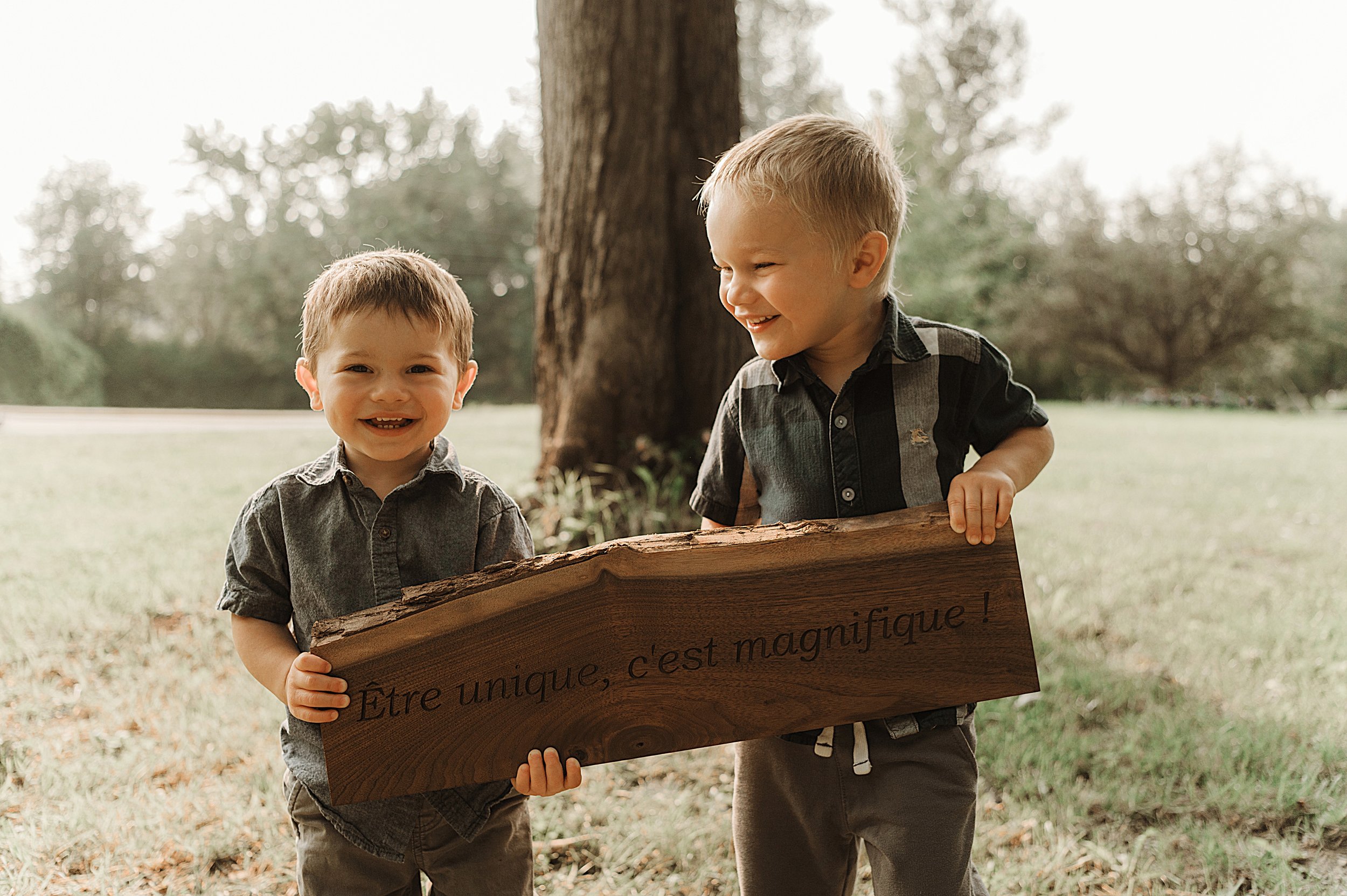 Two young boys outdoors near a tree, smiling and holding a wooden sign with French writing, wearing casual clothes.