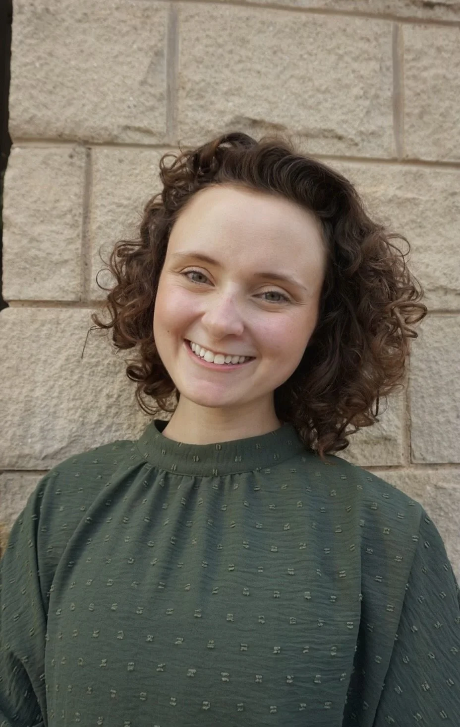 A young woman with curly brown hair smiling against a beige brick wall, wearing a green textured shirt.