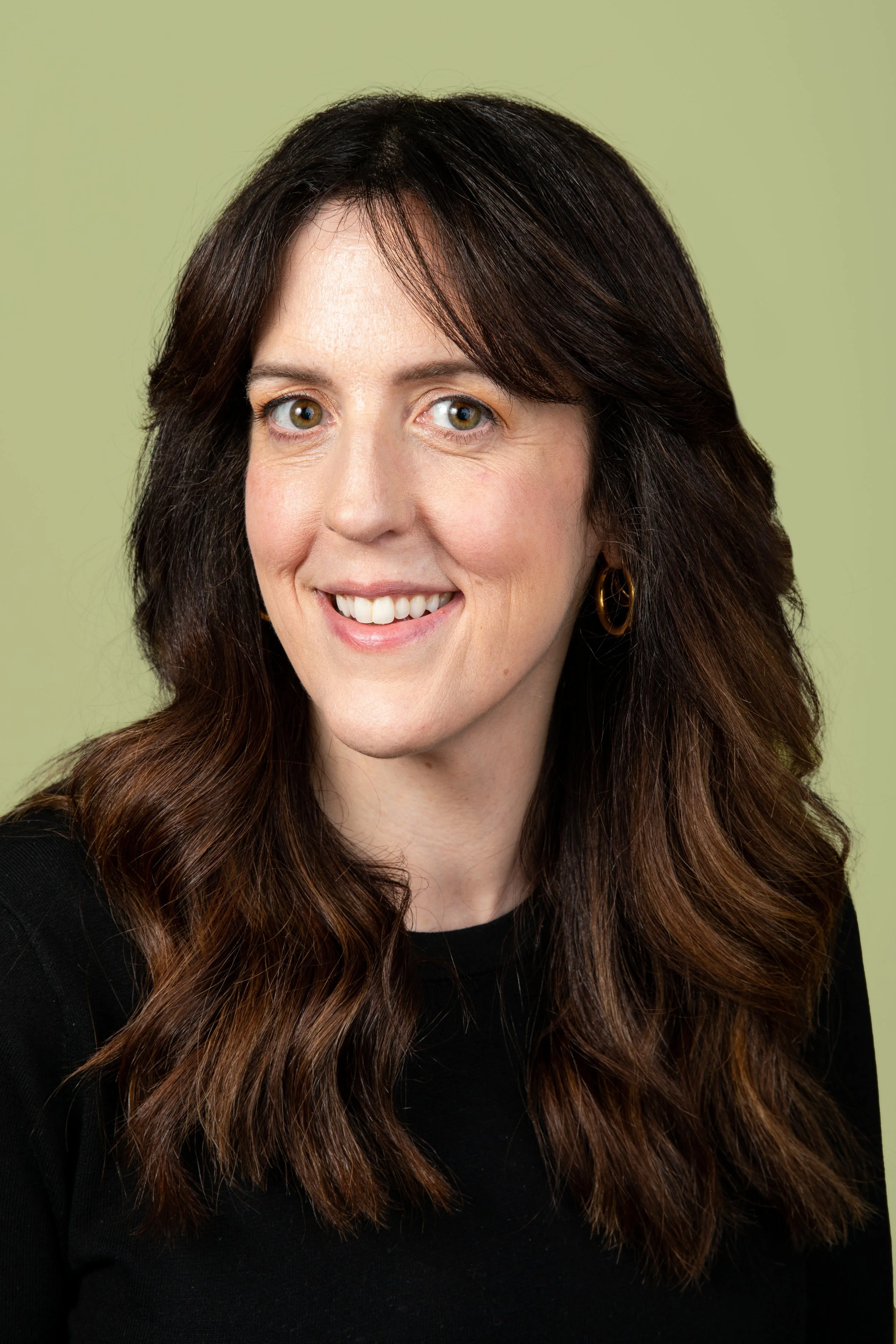 A woman with long wavy brown hair, light skin, brown eyes, wearing a black top and gold hoop earrings, smiling against a light green background.