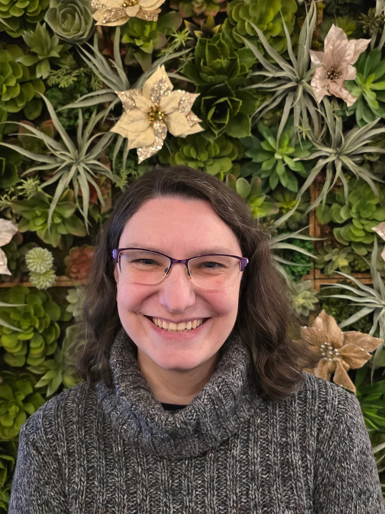 A woman with glasses smiling in front of a green foliage wall decorated with Christmas poinsettias and gold accents.