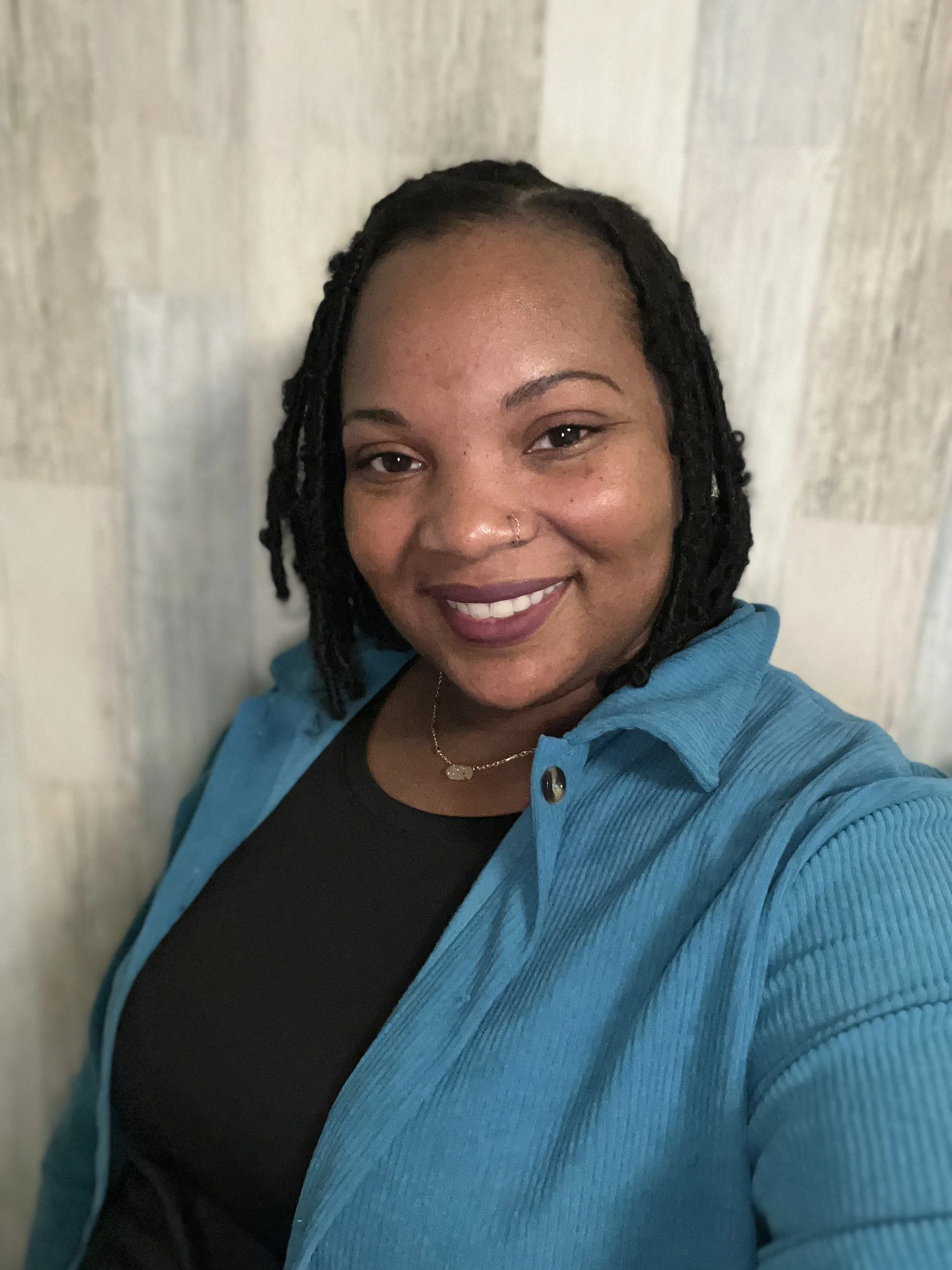 Close-up of a woman with shoulder-length dreadlocks, wearing a black shirt and a blue corduroy jacket, smiling and standing against a light wooden background.