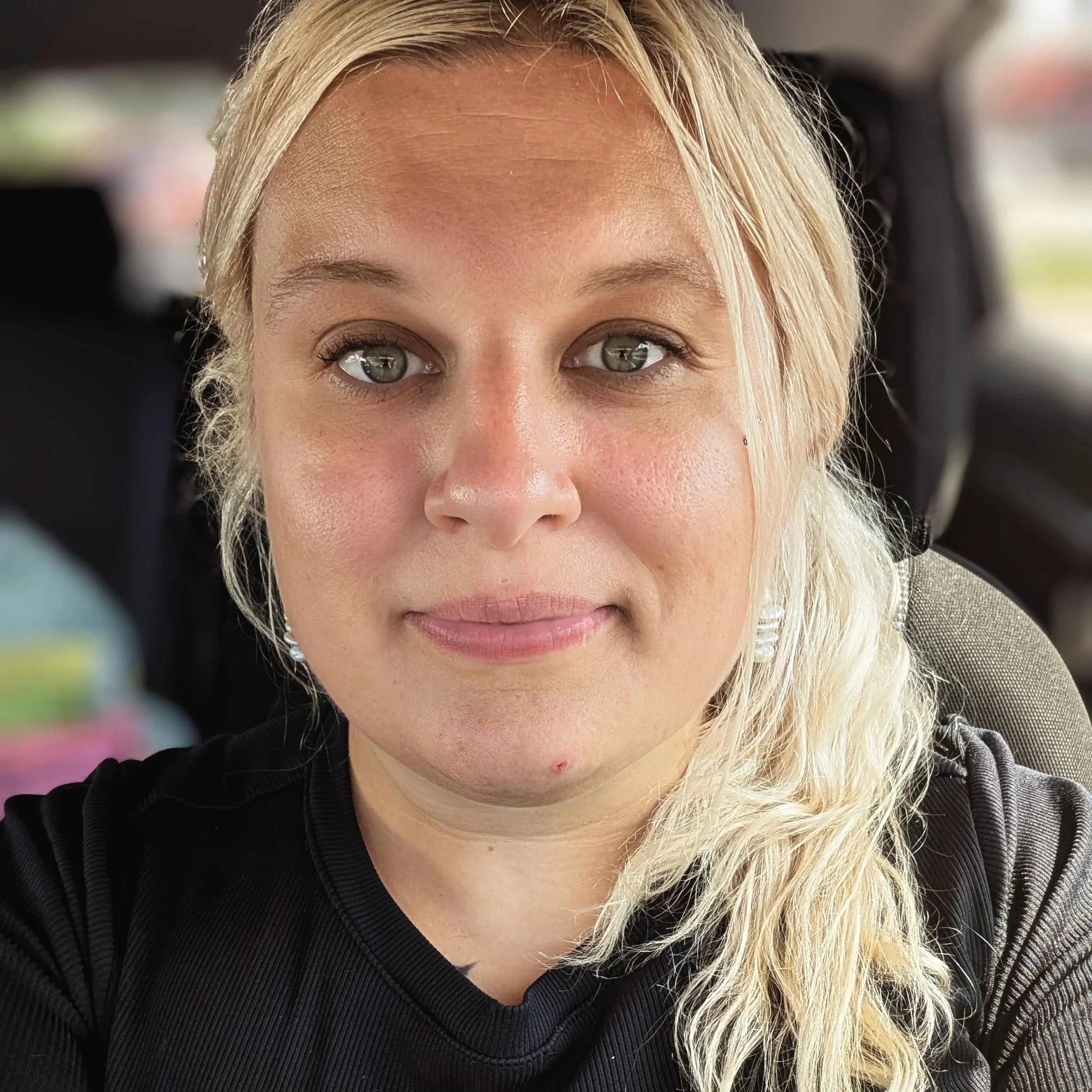 Close-up of a blonde woman with green eyes, wearing a black sweater, inside a vehicle.