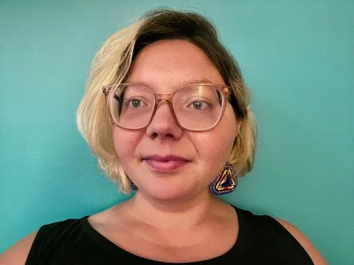A woman with short blonde hair, glasses, and a colorful earring, wearing a black top, standing against a teal background.
