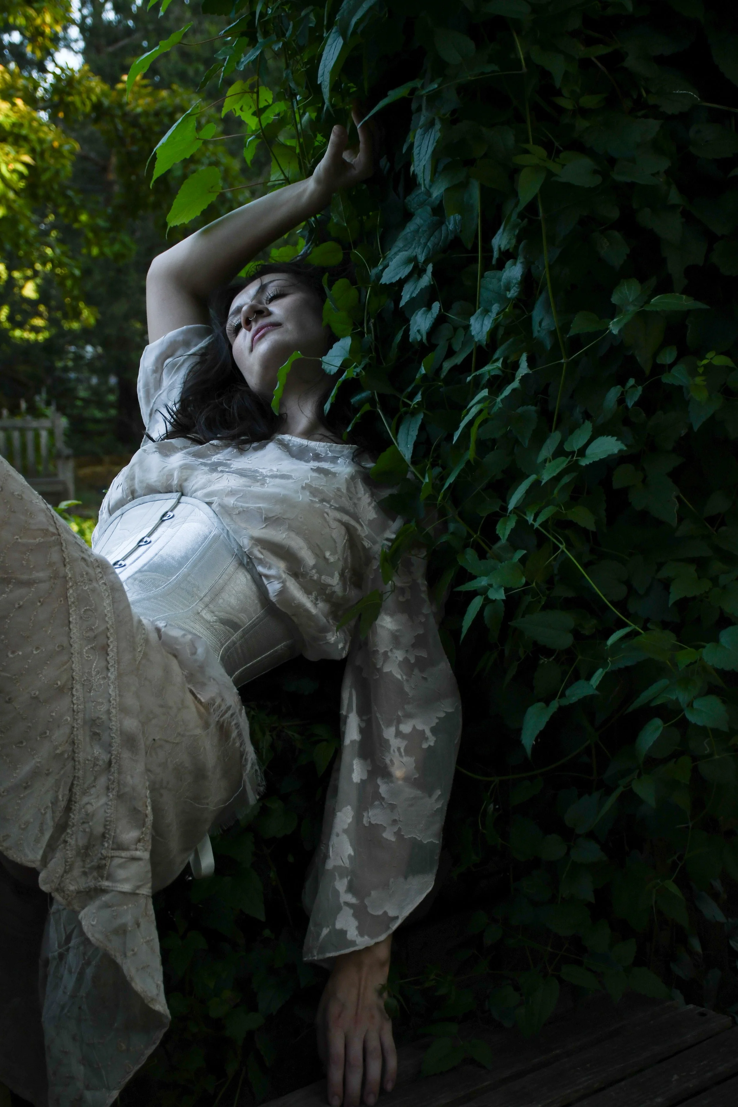 Woman with dark hair and a septum piercing lying on her side in a garden surrounded by green leaves, dressed in an ornate, light-colored dress with lace and embroidery details.