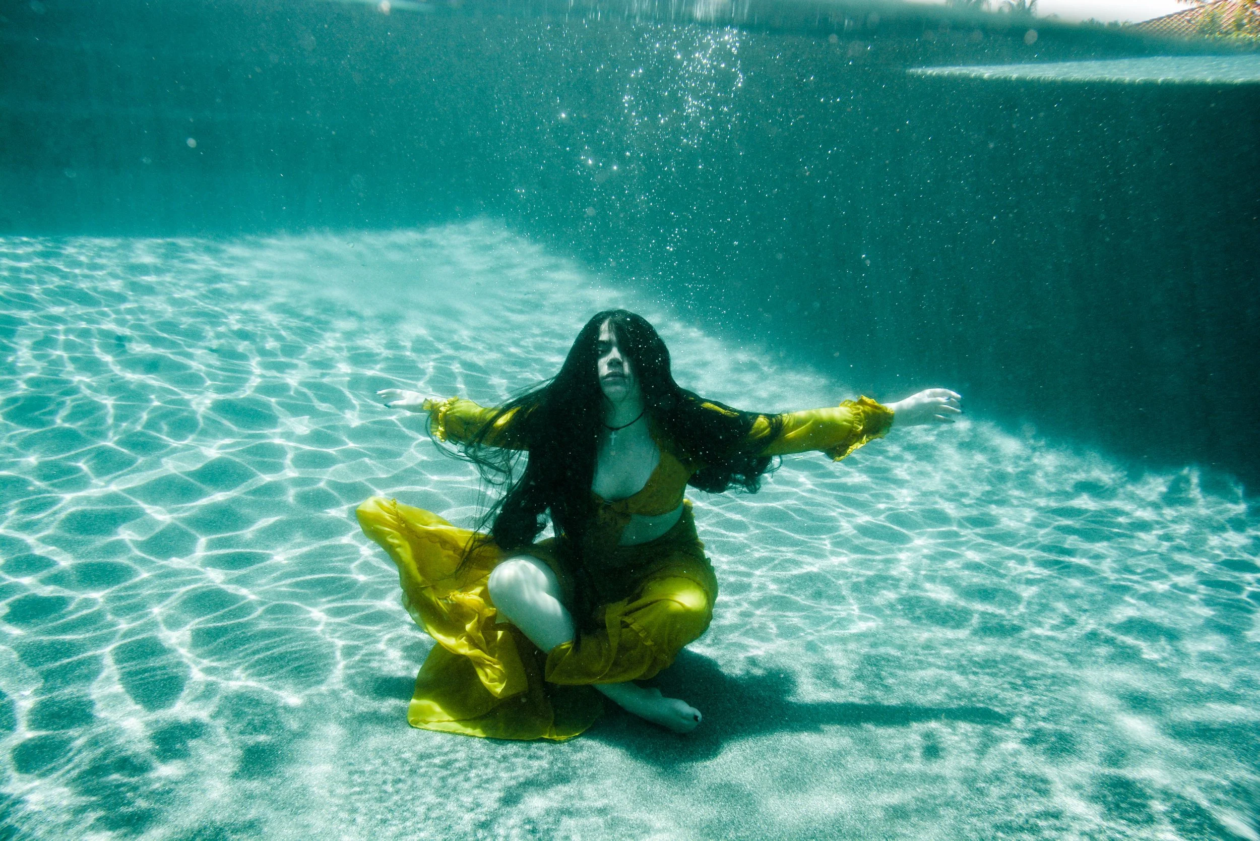A woman dressed in yellow raincoat and rain boots underwater in a swimming pool, sitting with legs crossed and arms stretched out to the sides.