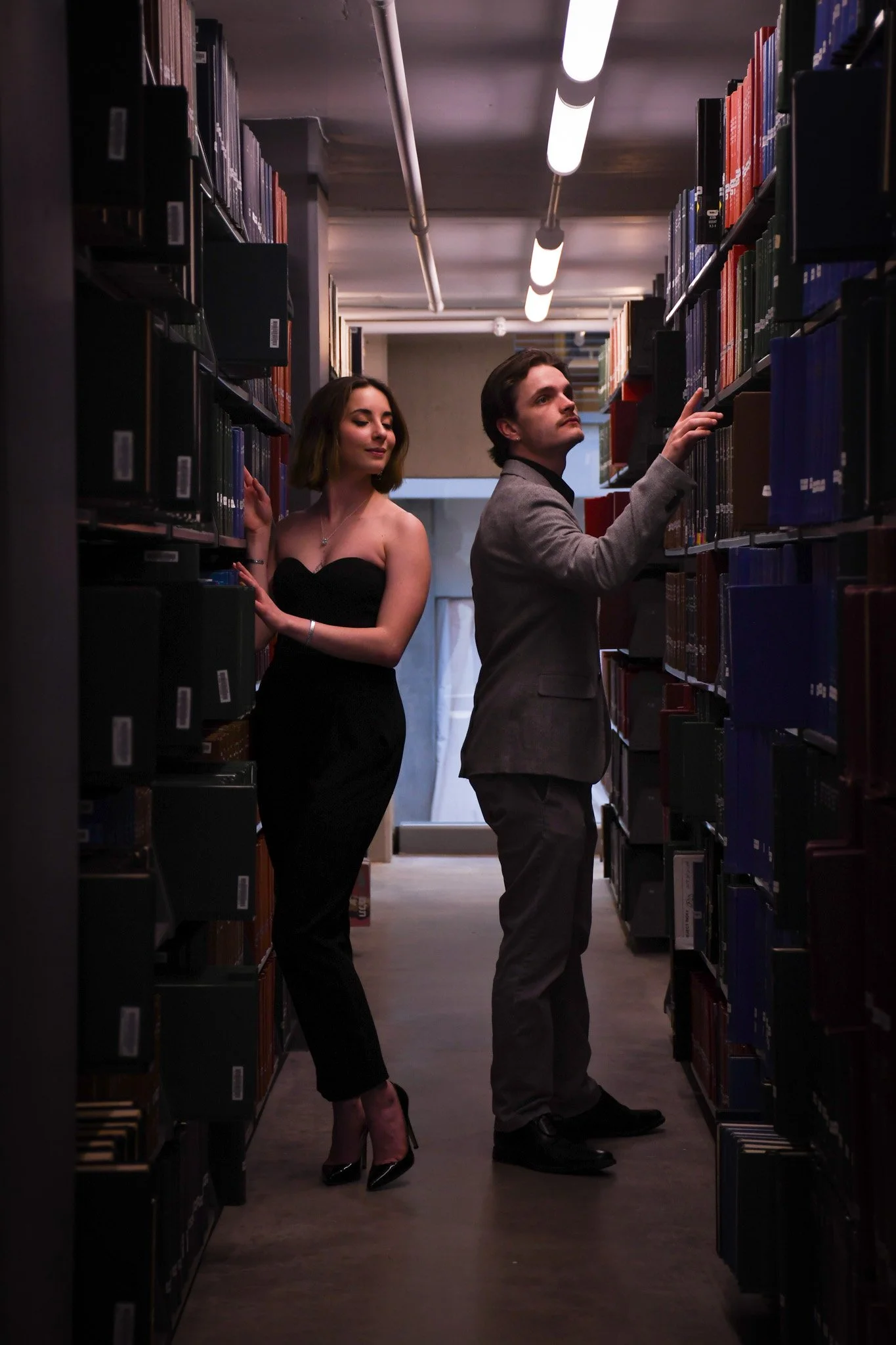 A man and woman in formal attire are standing in a library aisle, with the woman on the left wearing a strapless black dress and high heels, and the man on the right in a gray suit, examining books on the shelves.