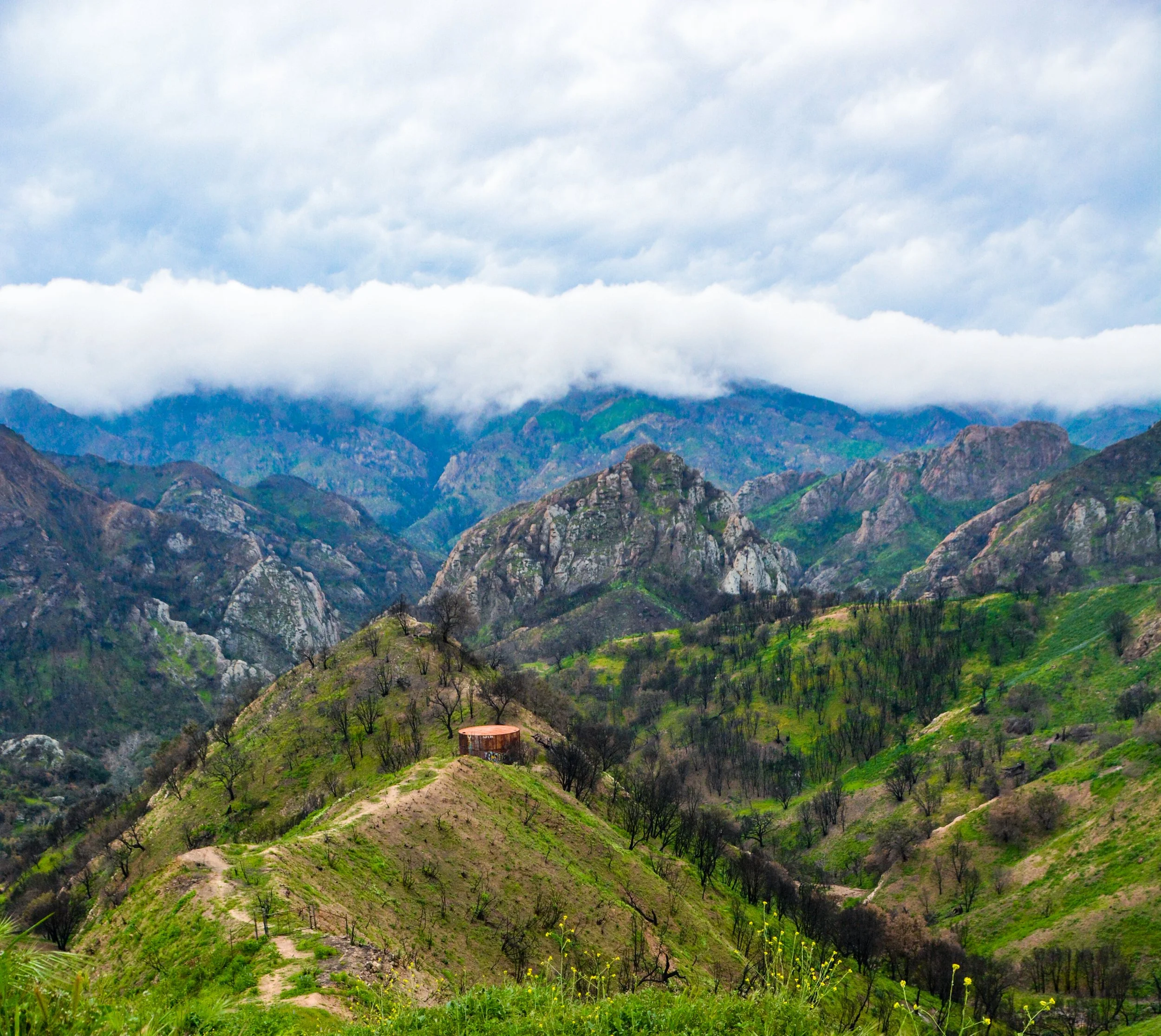 Mountain landscape with green hills, rocks, sparse trees, and a cloudy sky.
