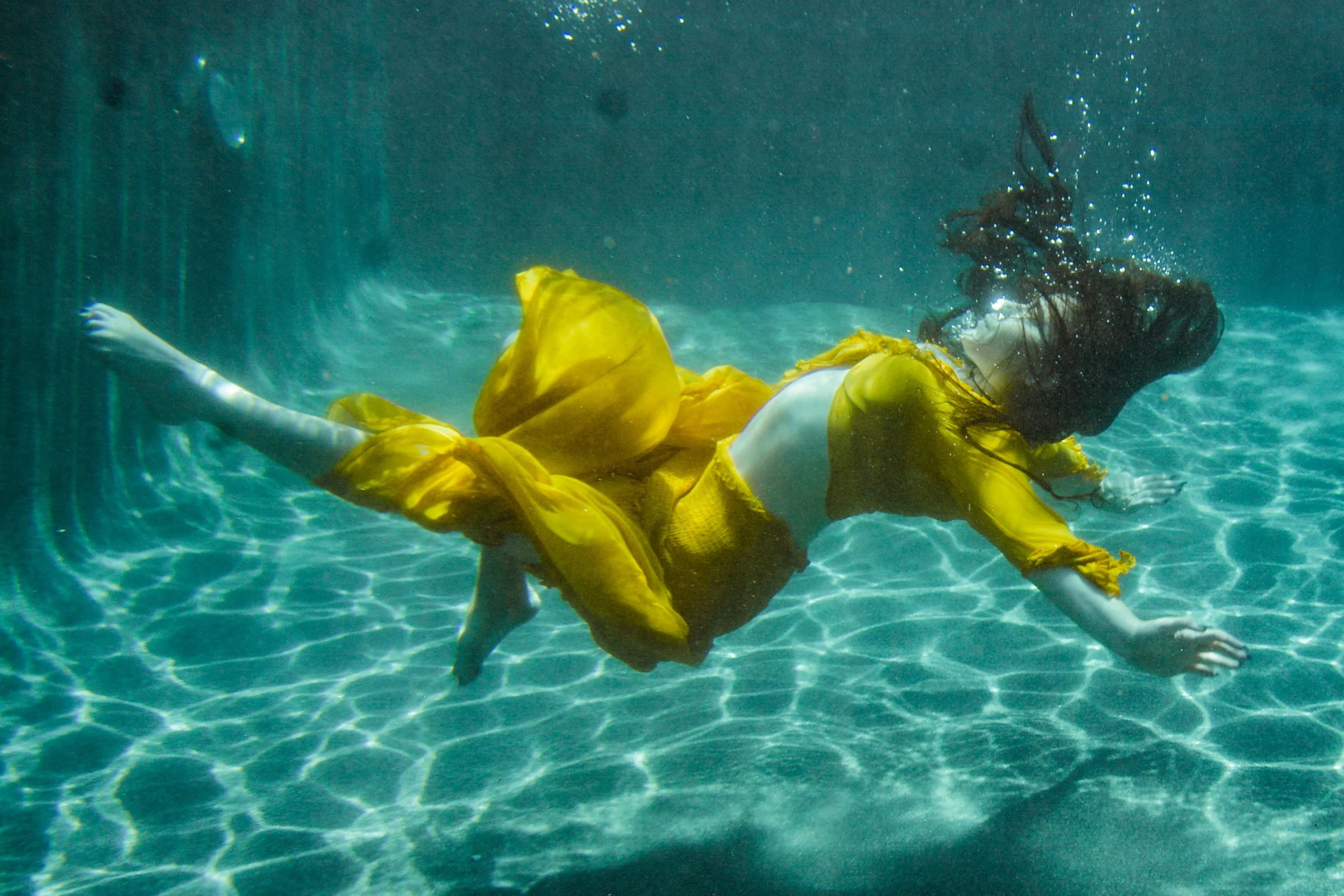 A person in a yellow dress or top is underwater, lying on the pool floor with arms and legs extended. Their dark hair floats freely around their head, and the water is clear with light reflections on the surface.