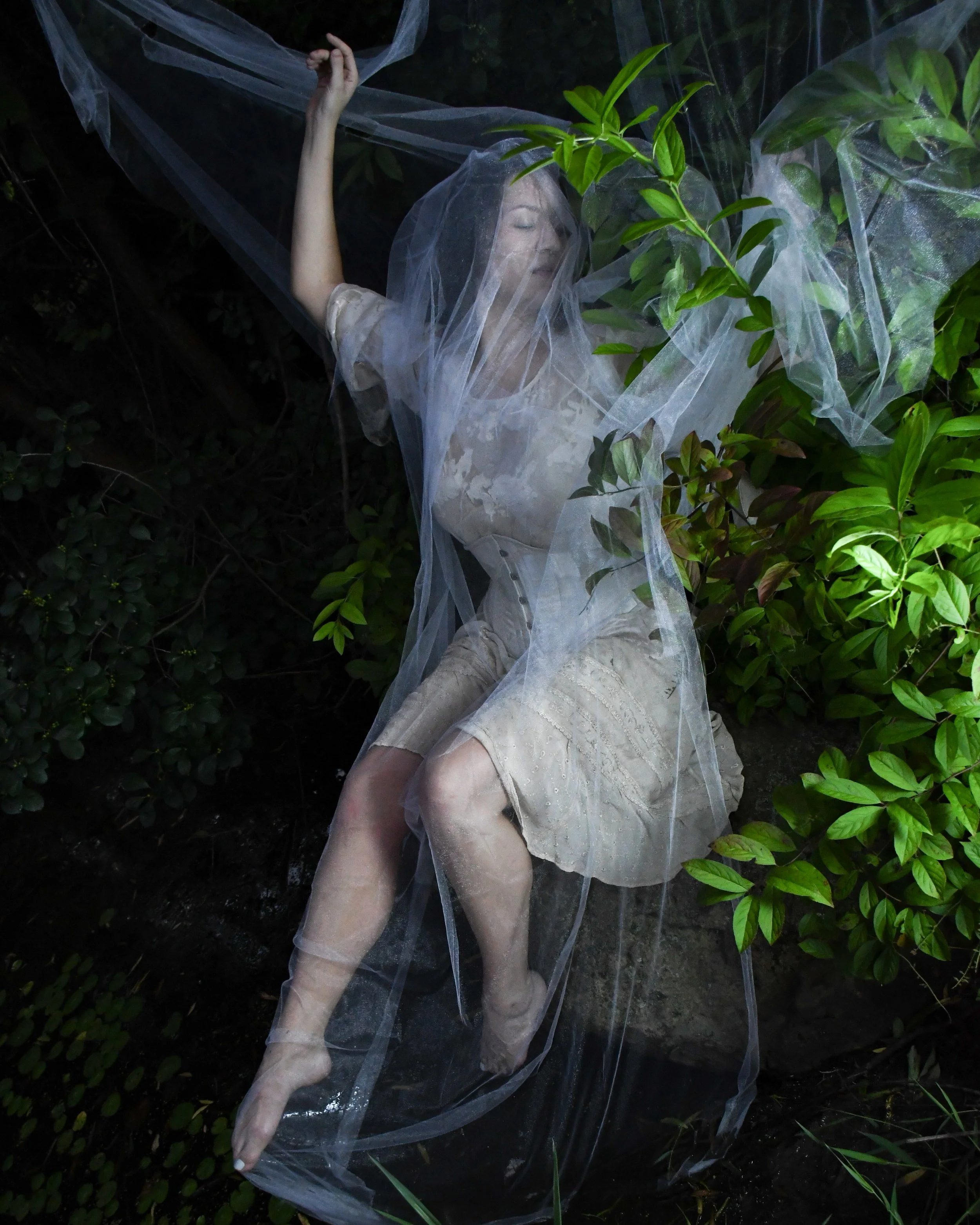 A woman with closed eyes and a serene expression, lying among greenery, with a sheer fabric draped over her head and body, creating a dreamlike scene.