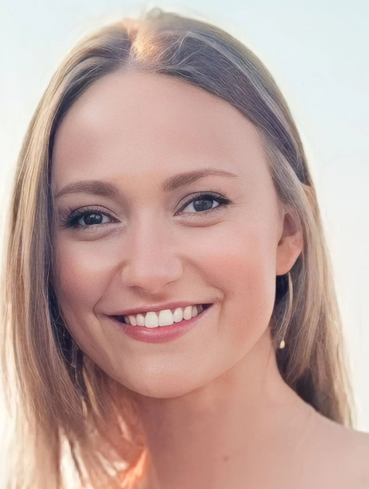 Close-up of a young woman with long blonde hair, smiling, showing her teeth, with bright eyes and a light background.