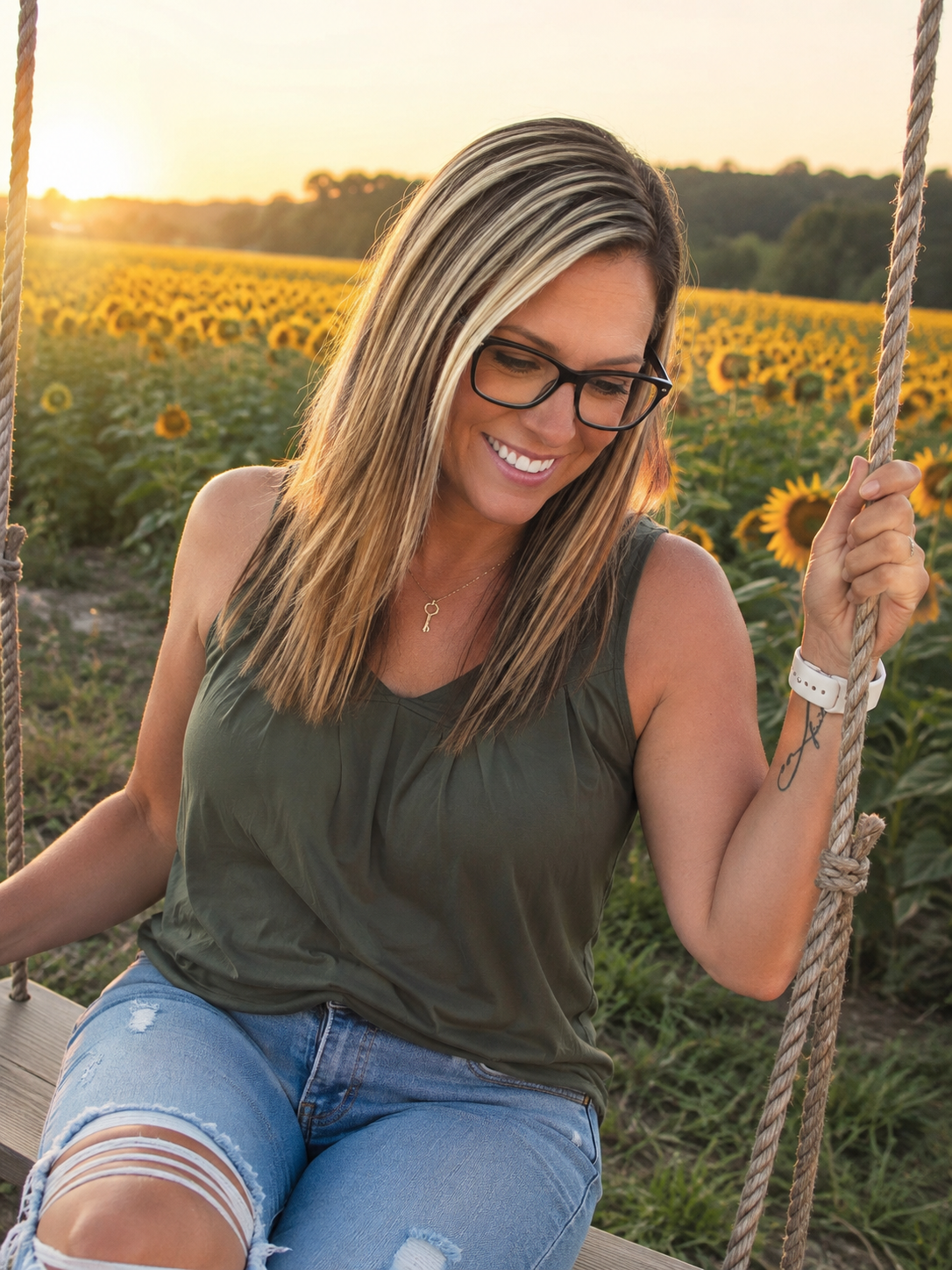 A woman with long hair, wearing glasses, a sleeveless top, and ripped jeans, smiling on a swing in a sunflower field at sunset.