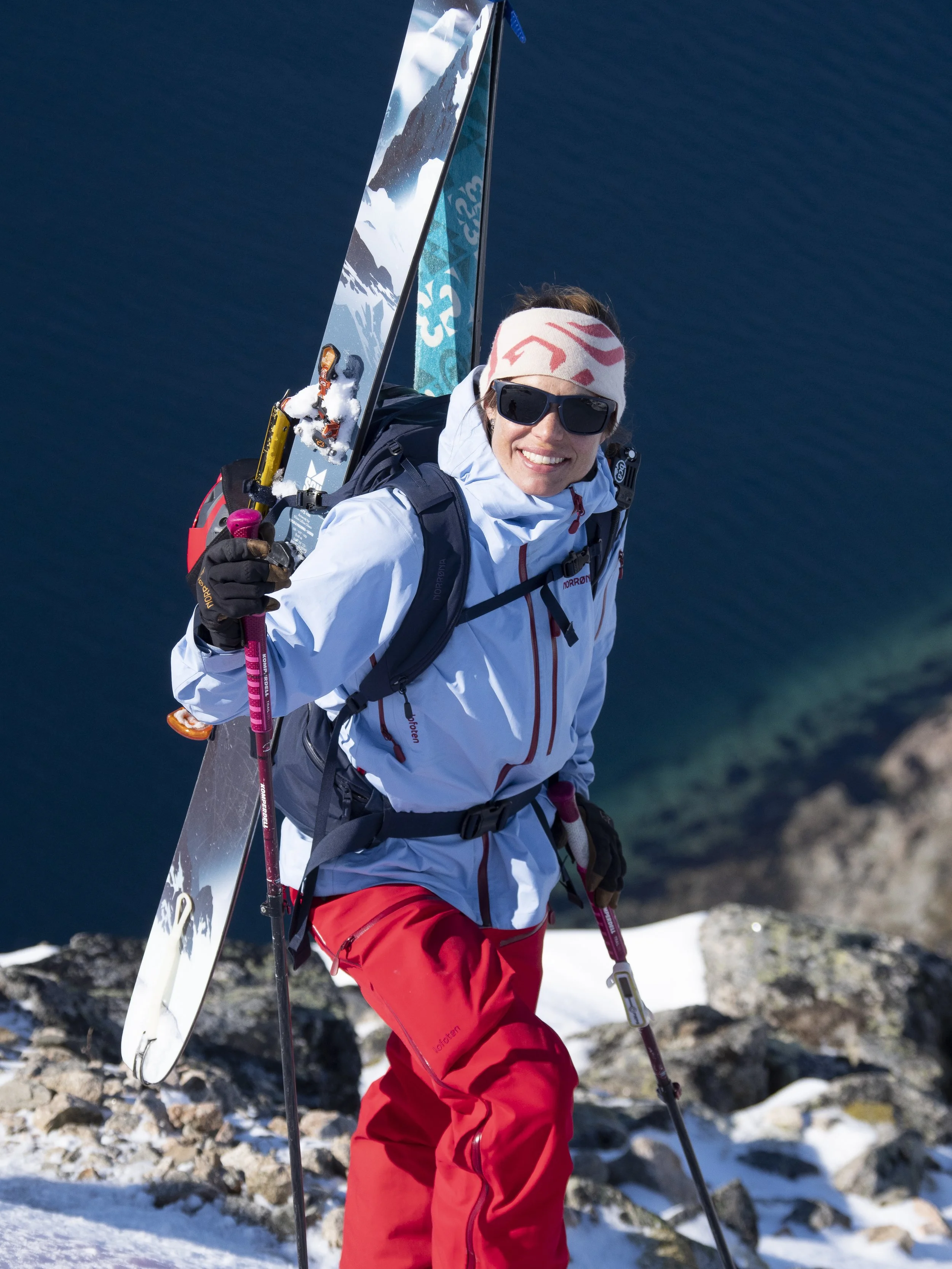 A person dressed in winter outdoor gear, holding ski poles, hiking on rocky snow-covered terrain near water, with a ski and mountains visible.
