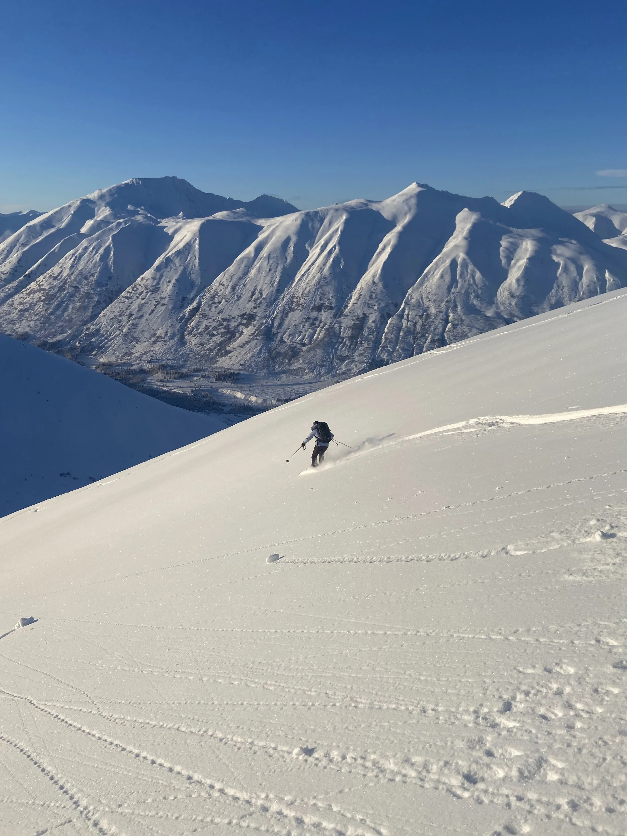 A person skiing on a snowy mountain slope with a backdrop of snow-covered mountains under a clear blue sky.