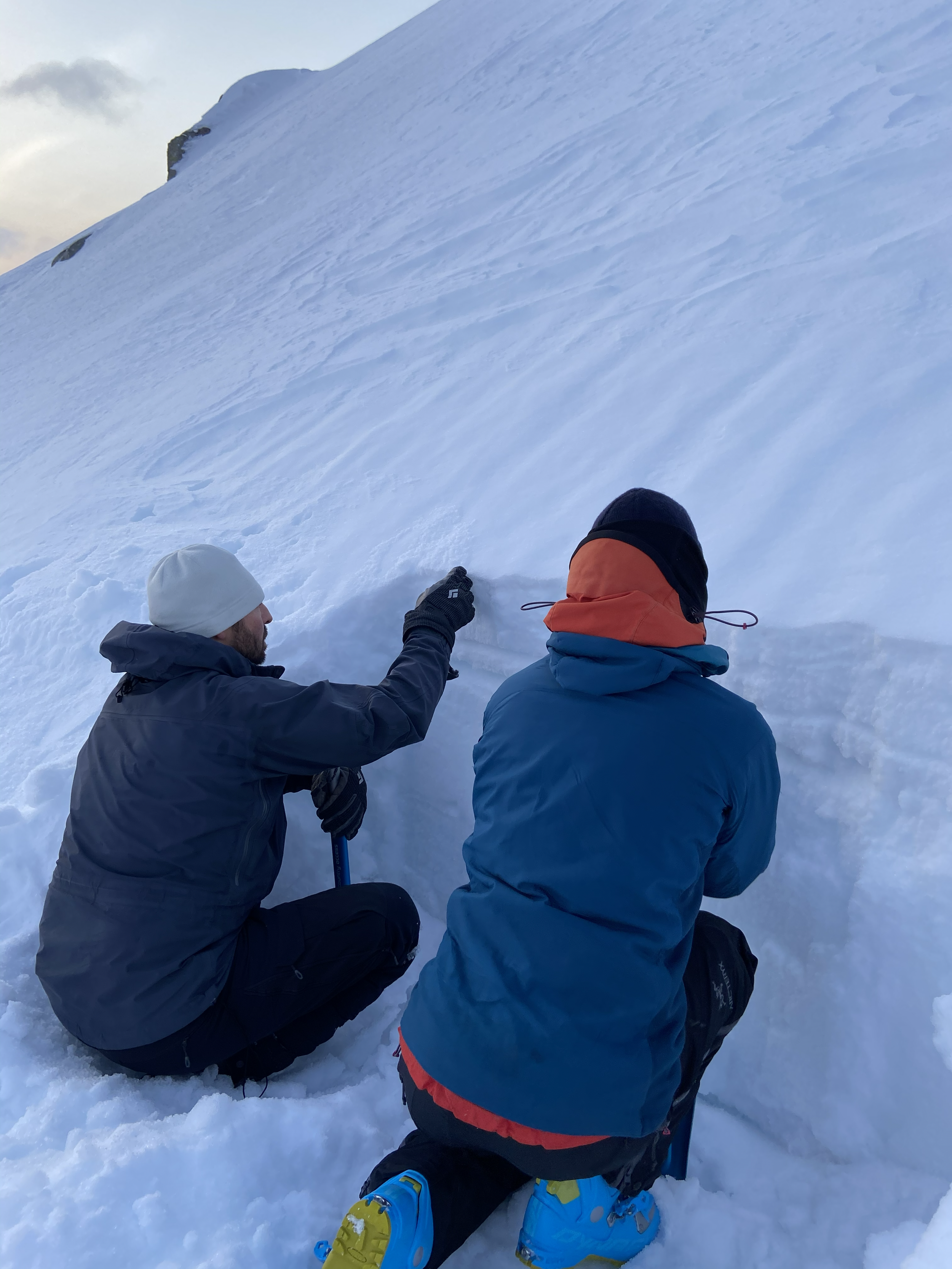 Two climbers or mountaineers resting or preparing on a snow-covered mountain slope, wearing winter gear and helmets, with one person pointing or adjusting equipment.