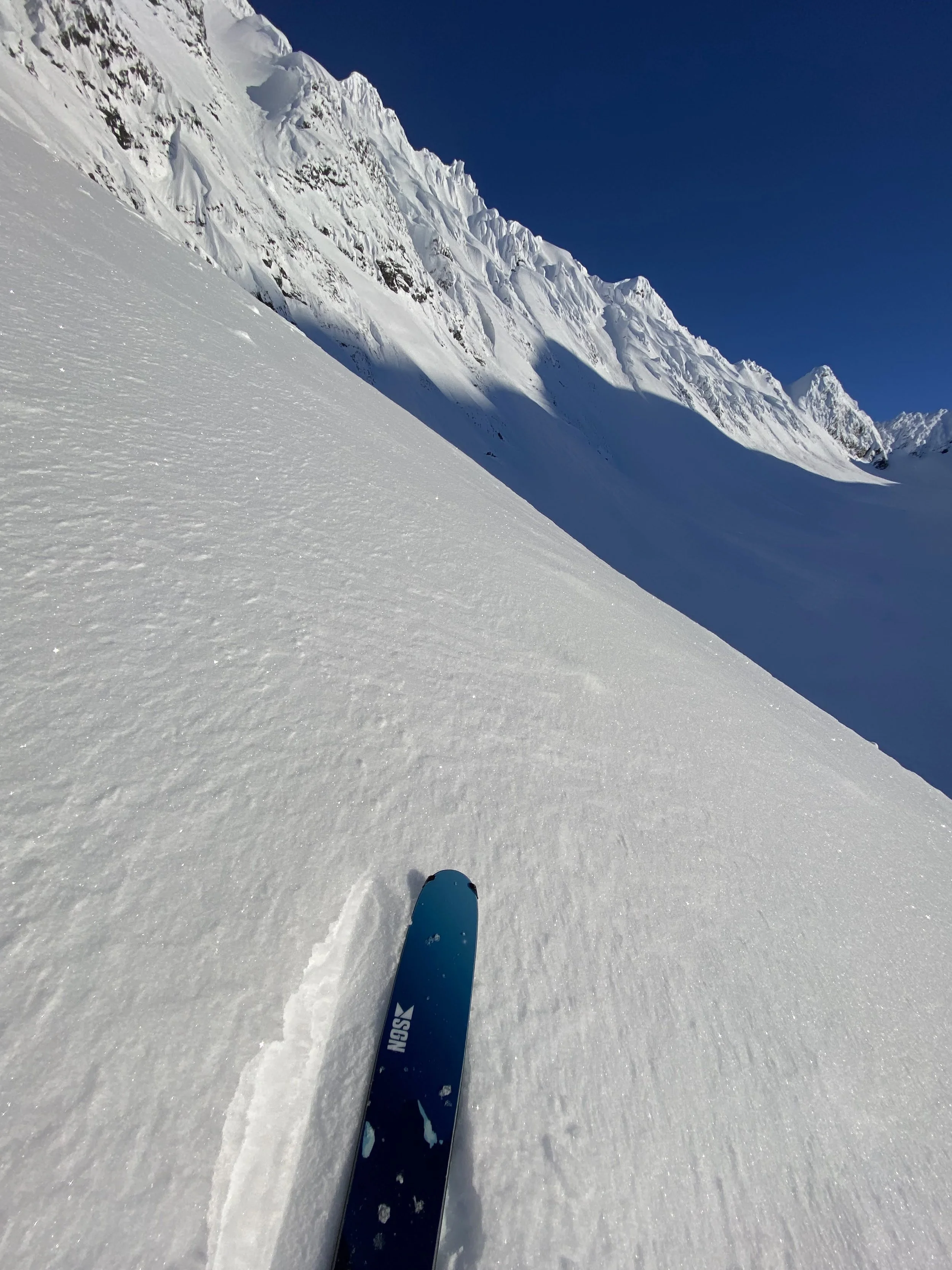 A skier's perspective traveling down a snow-covered slope with a large snowy mountain range in the background under a clear blue sky.