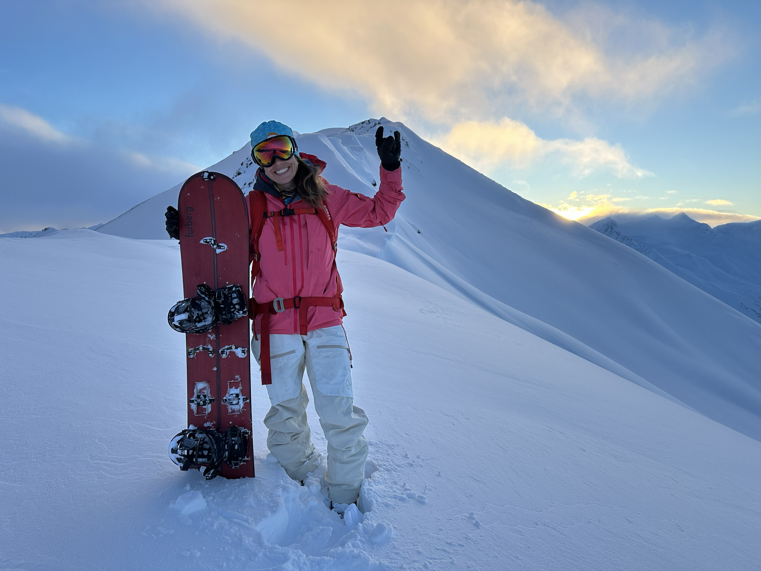 A woman in winter gear, wearing a pink jacket, white pants, and ski goggles, stands on snowy mountain terrain holding a snowboard and making a peace sign with her hand, with snow-covered peaks and a partly cloudy sky in the background.