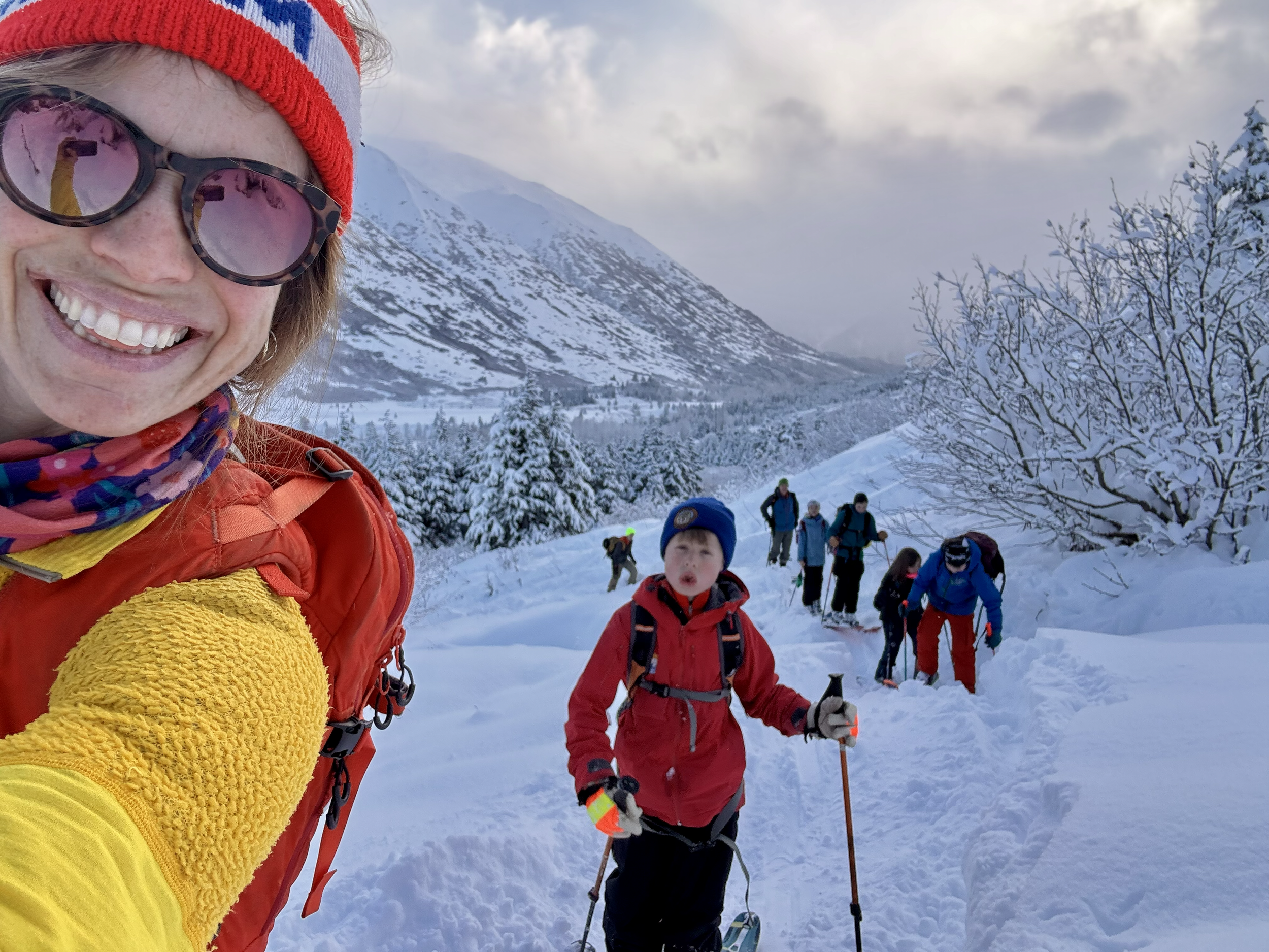 A woman smiling in sunglasses and a red, white, and blue beanie taking a selfie in snowy mountains. Behind her, a group of children and adults are snowshoeing or hiking with snowy trees and mountains in the background.