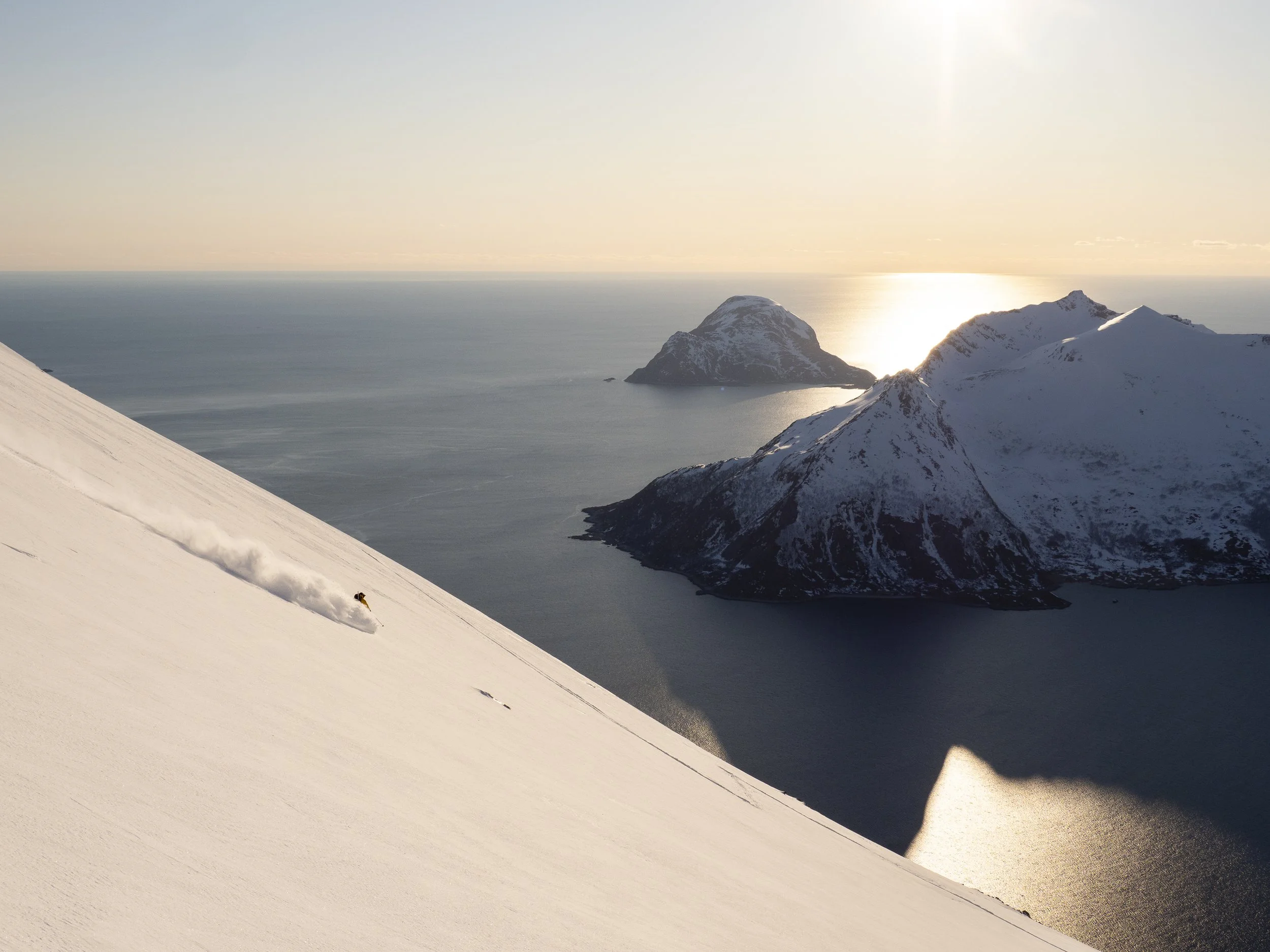 A skier descending a snow-covered mountain slope, with snow-capped mountains and ocean in the background during sunset.