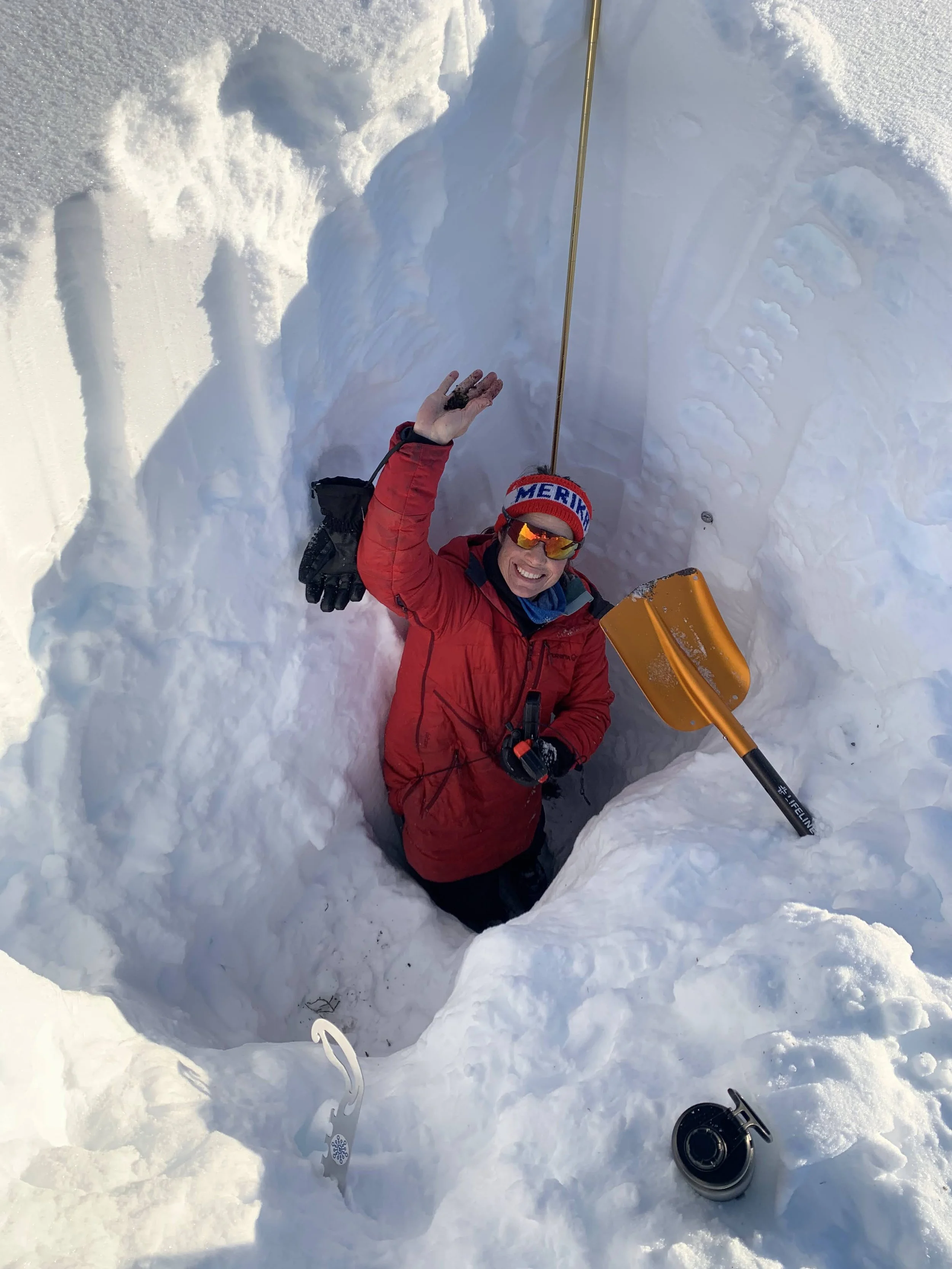 A person in a red jacket, wearing sunglasses and a hat, is lying on snow in a small snow tunnel with a smile, holding a yellow snow shovel, with a fishing reel and snow measuring tools nearby.