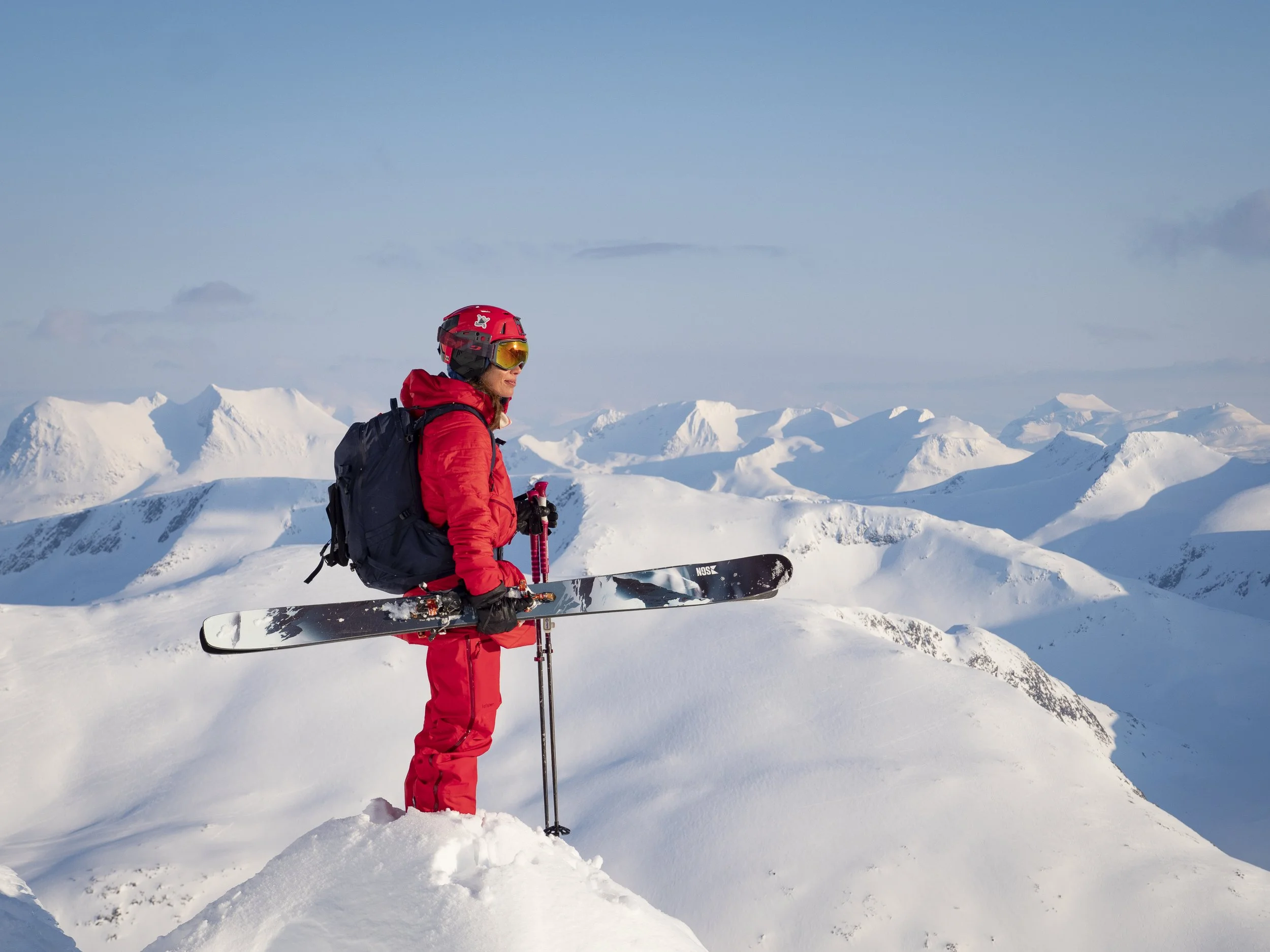 A person dressed in red winter gear, wearing a helmet and skiing goggles, stands on snow-covered terrain with snow-capped mountains in the background.