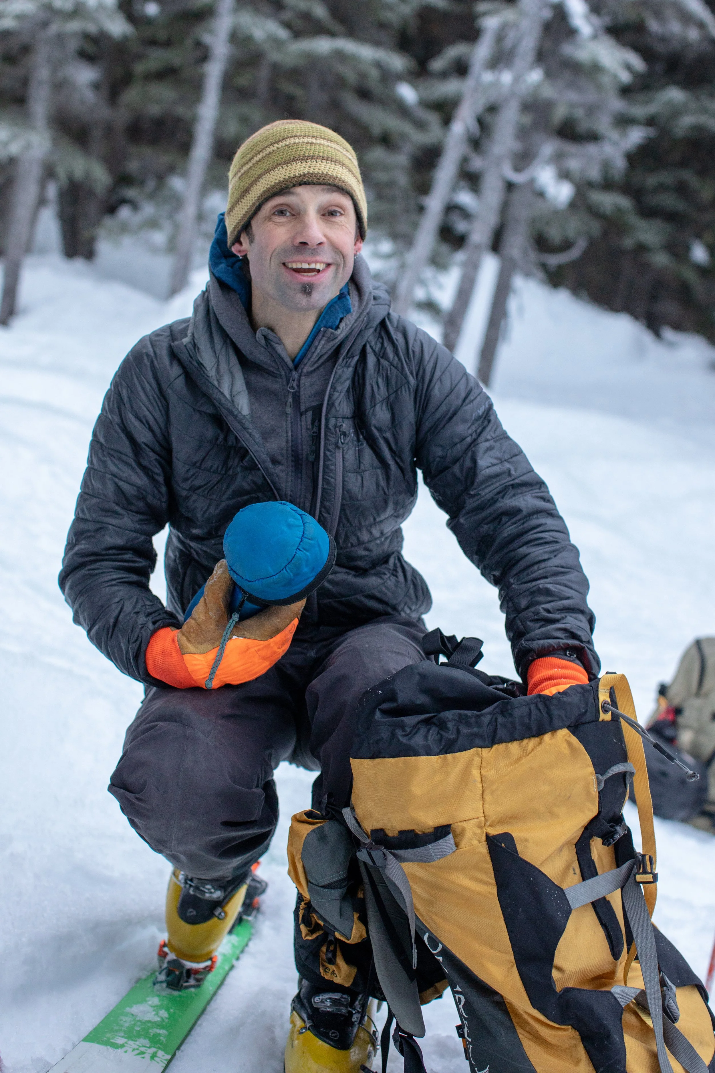 A man with a green beanie and a black winter jacket sitting on snow, smiling while holding a blue thermos. He is in a snowy forest and wearing ski boots with skis on his feet. There are trees in the background.