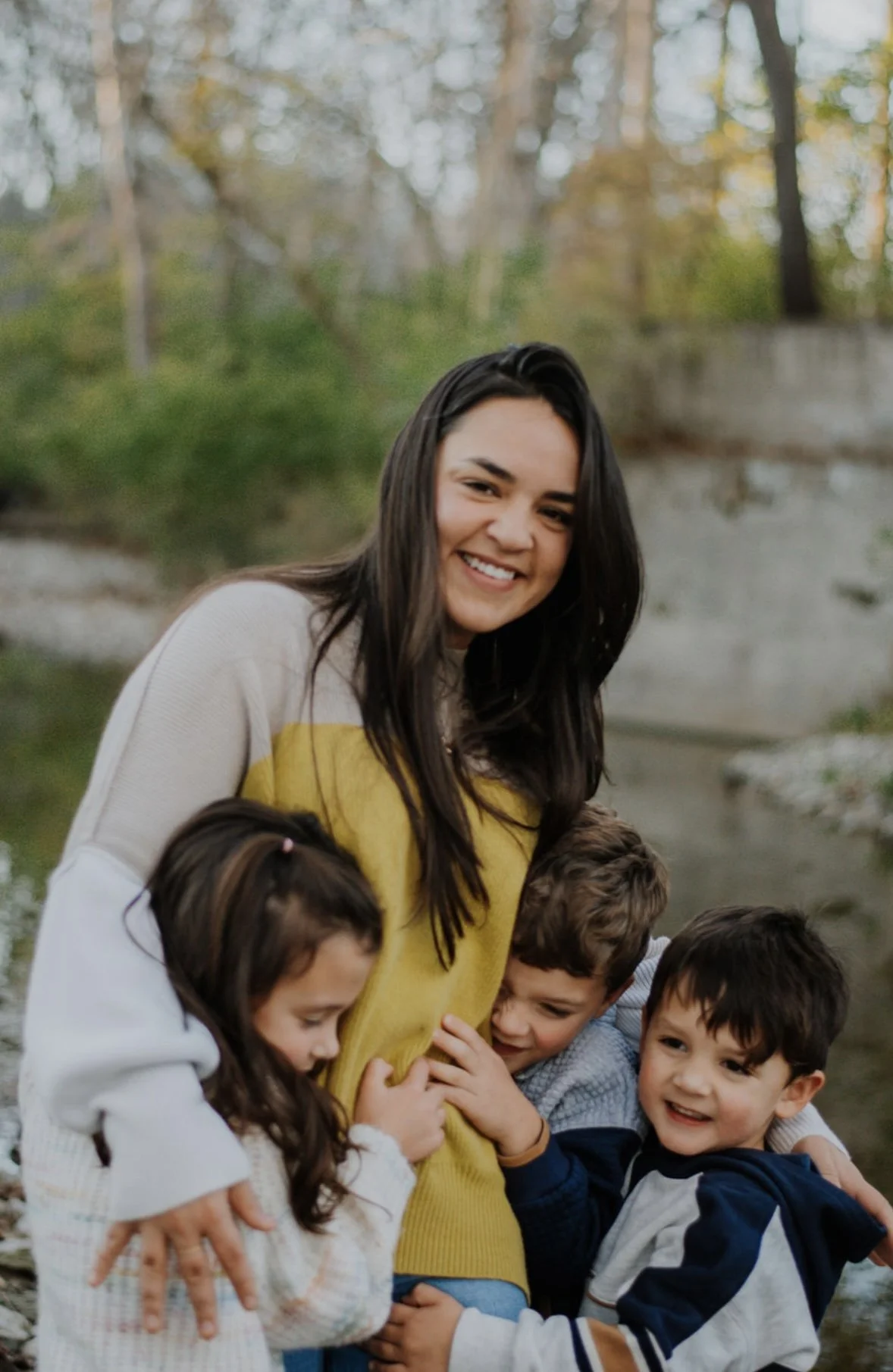 A smiling young woman with long dark hair surrounded by children hugging her outdoors near a river or creek, with trees and autumn foliage in the background.