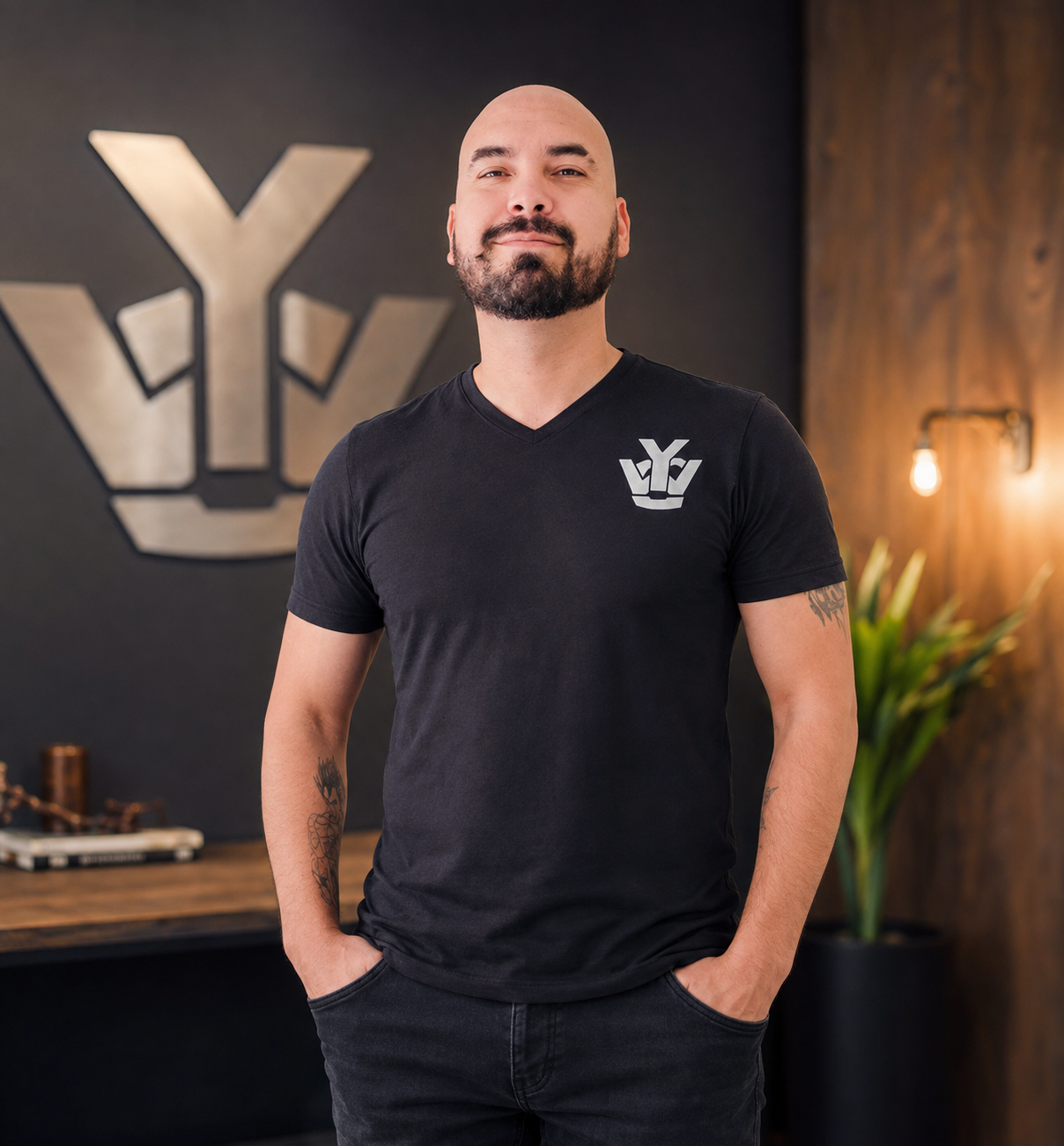 A confident man with a beard and tattoos wearing a black V-neck T-shirt with a logo on the chest, standing indoors in front of a large logo and warm lighting.