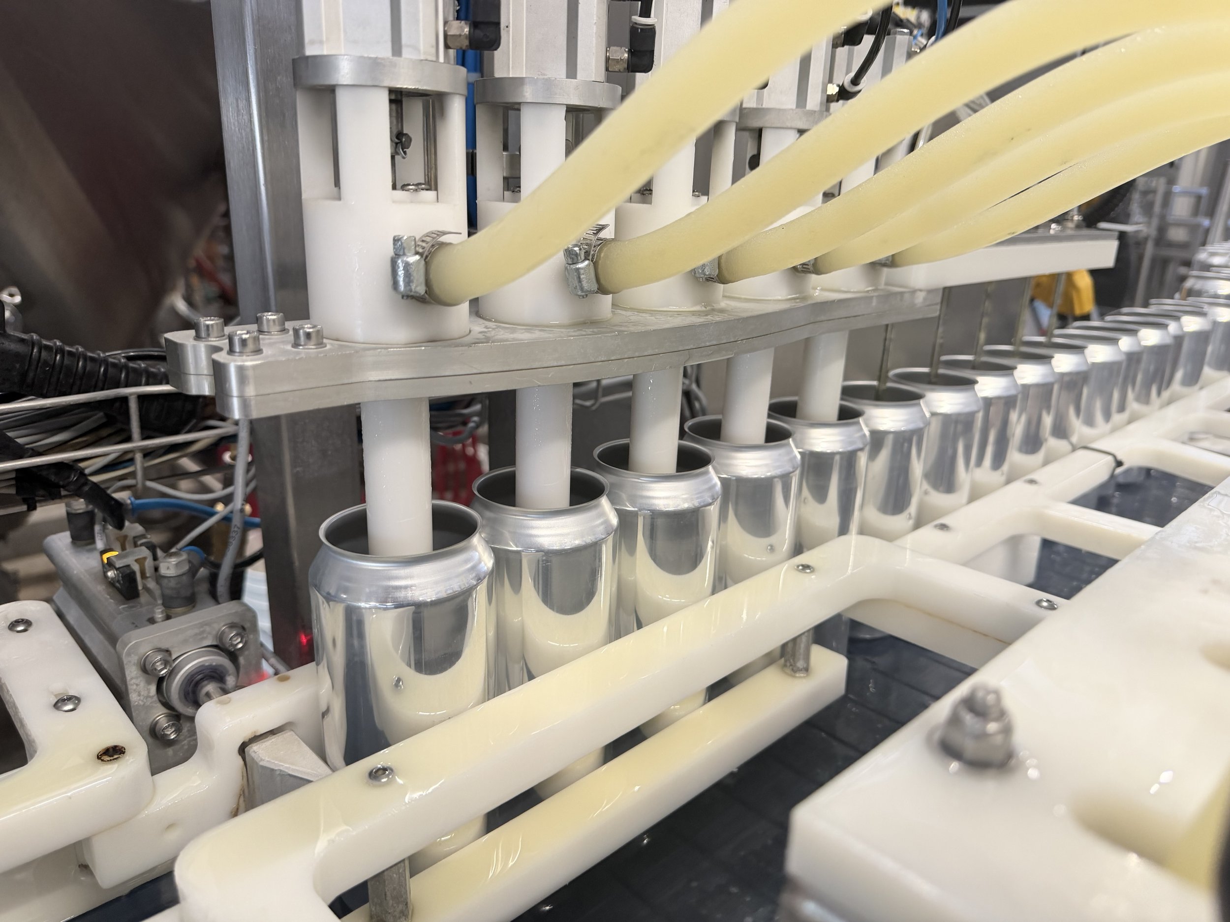 Close-up of a manufacturing process with yellow flexible tubes, metal fittings, and cylindrical metal containers, possibly filling or packaging liquid or food products.