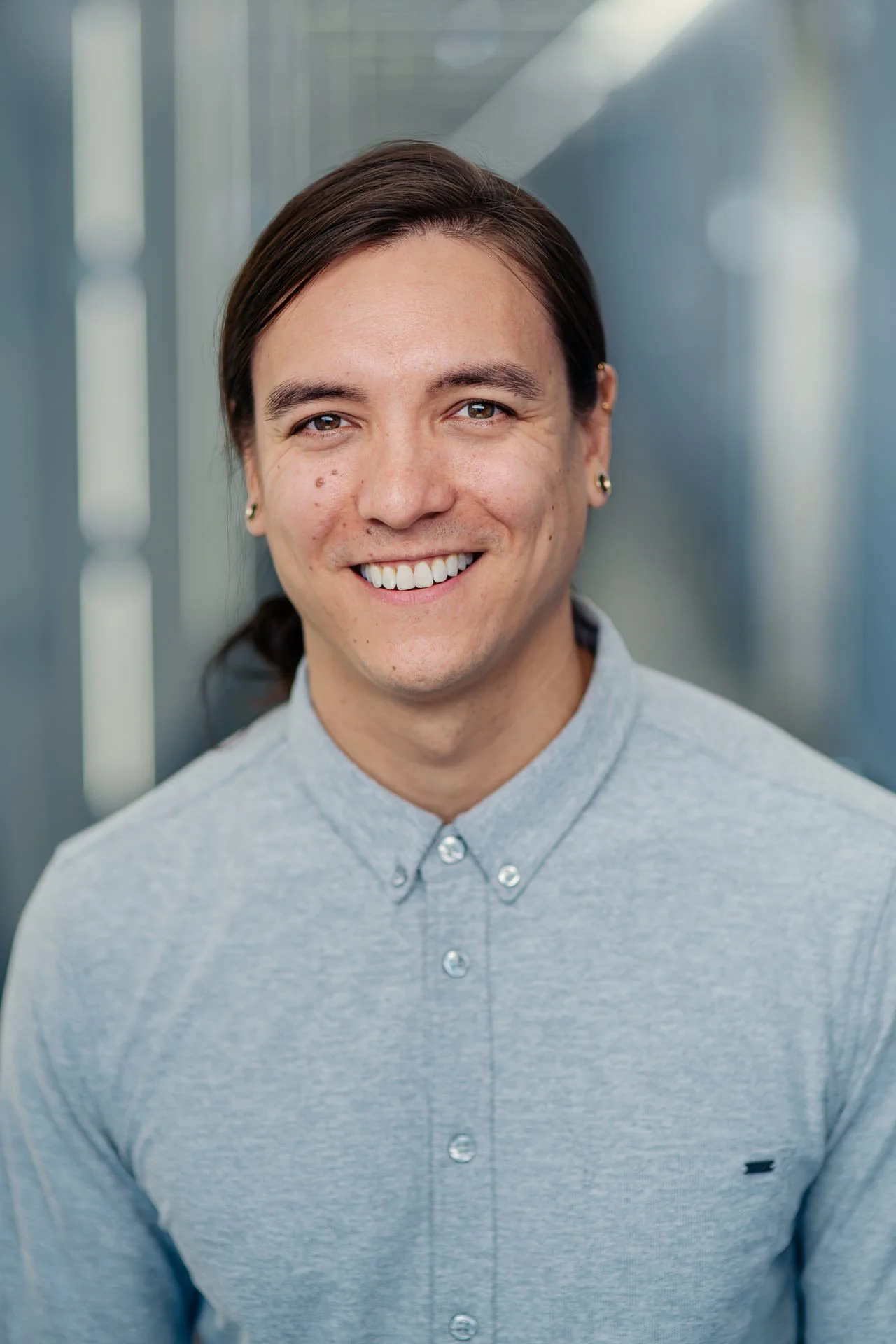 Close-up portrait of a smiling man with brown hair, brown eyes, and fair skin, wearing earrings and a light gray collared shirt, standing in an indoor setting with blurred background. That man is the lab PI, Andrew