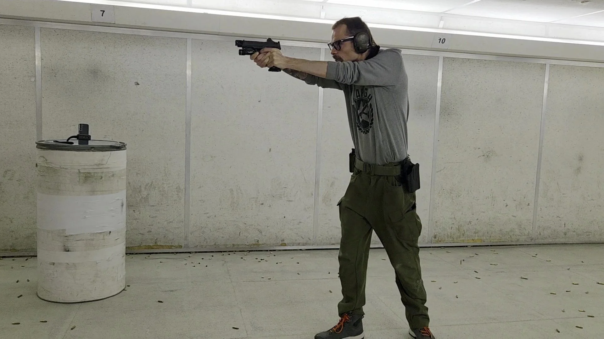 Man practicing handgun shooting indoors, aiming with a pistol while wearing hearing protection and glasses, standing in front of a wall with numbered lanes.