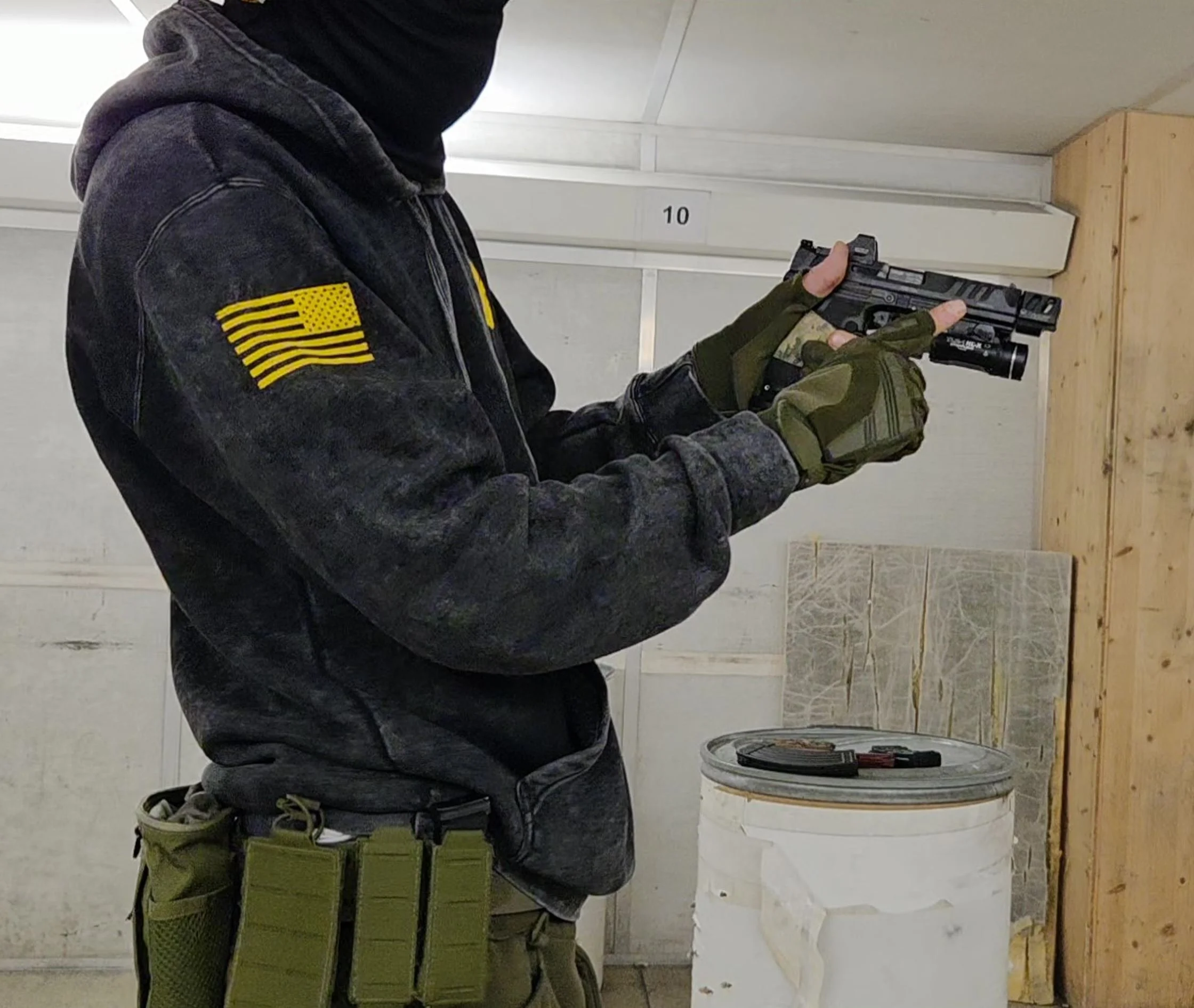 Person in black hoodie with yellow American flag patch, camo gloves, green tactical gear, and dark backpack, holding a handgun at an indoor shooting range. Wooden target stand and a white washing machine are visible in the background.