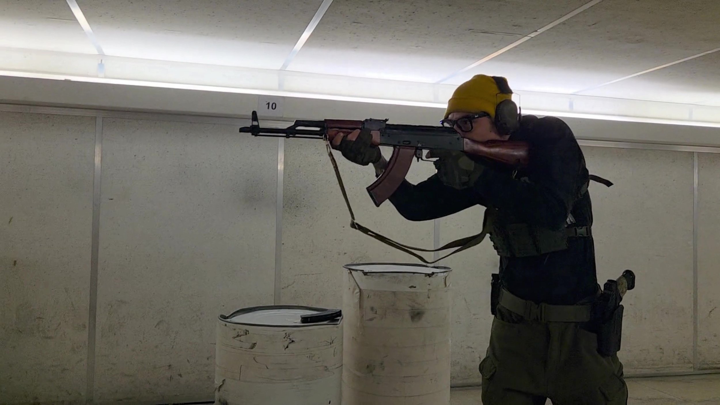 Person wearing tactical gear and ear protection aiming a rifle at an indoor shooting range.