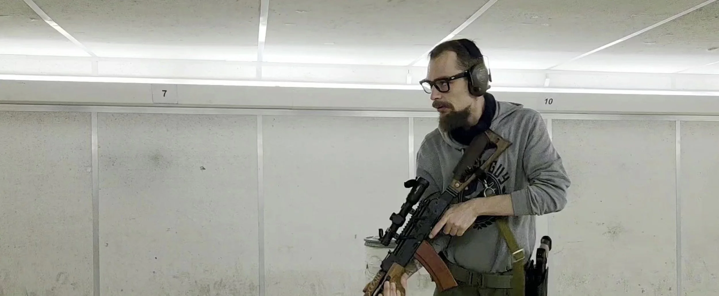 A man with glasses, a beard, and headphones, holding a rifle at a shooting range, standing in front of numbered targets.