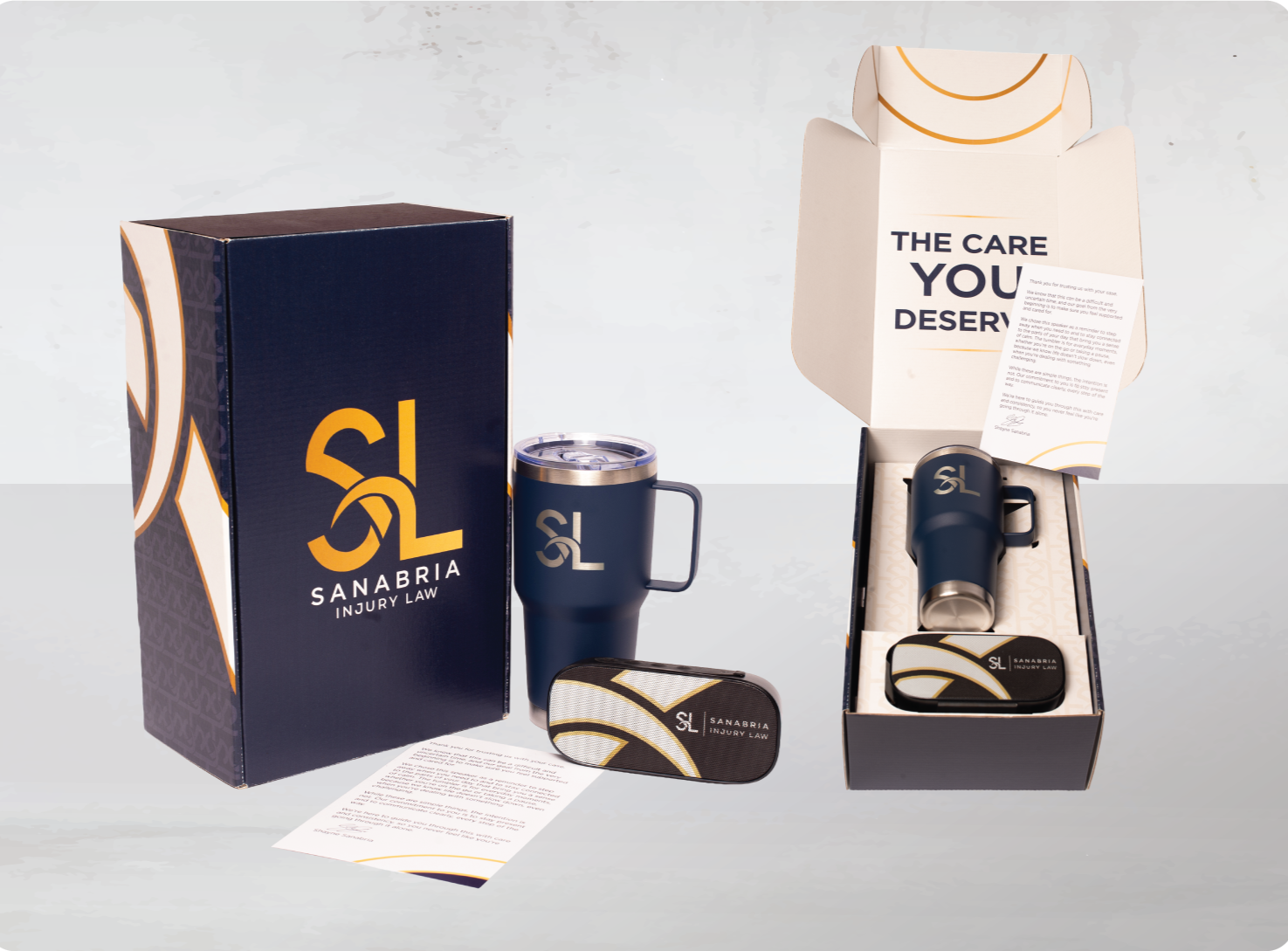 A Sanabria Injury Law branded gift set includes a dark blue insulated travel mug with a handle, a matching zippered case, a ceramic mug, a cardboard box, and a folded informational leaflet.