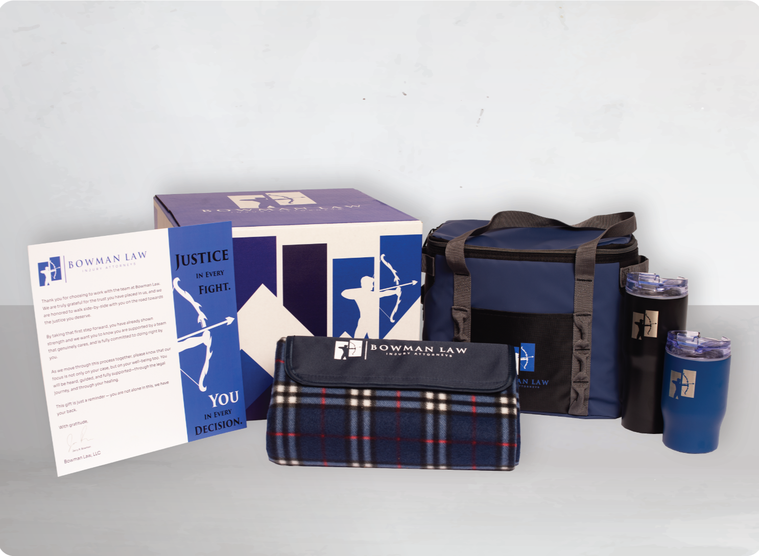 Bowman Law branded gift set including a box, a letter, a blanket, a lunch bag, and two travel mugs, all in navy blue with law firm logo and motivational messages.