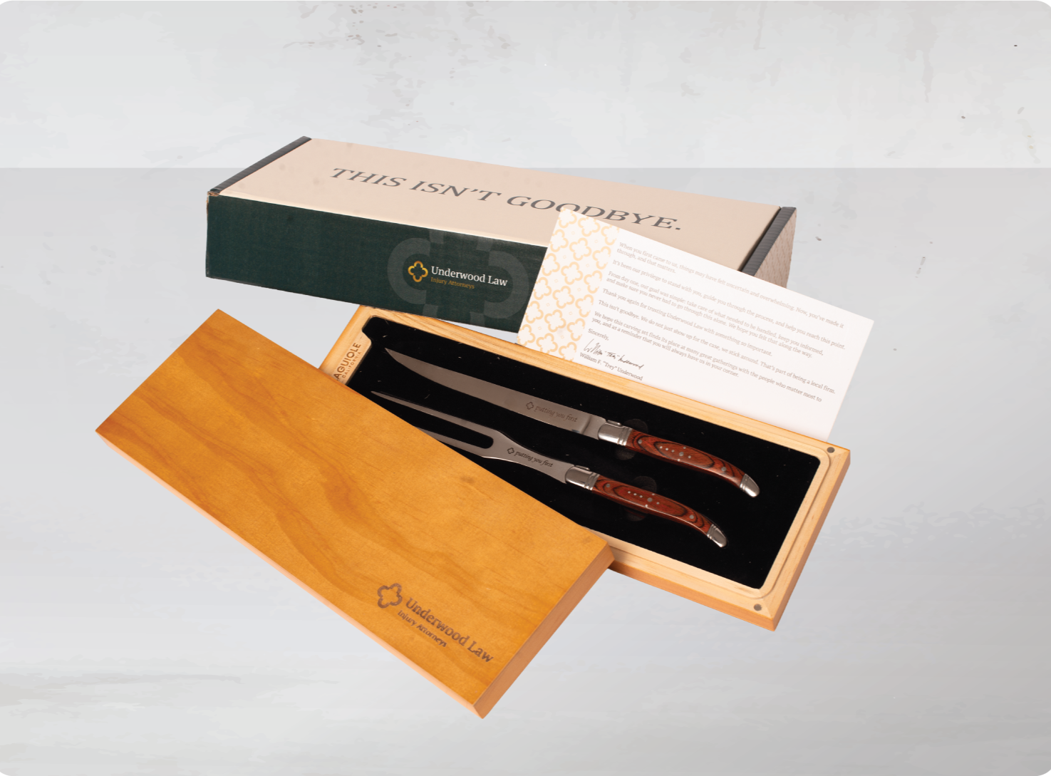 A gift set of two steak knives with wooden handles in a wooden box, with a white card, a black gift box labeled 'Underwood Law Injury Attorneys', and a paper insert inside the box.