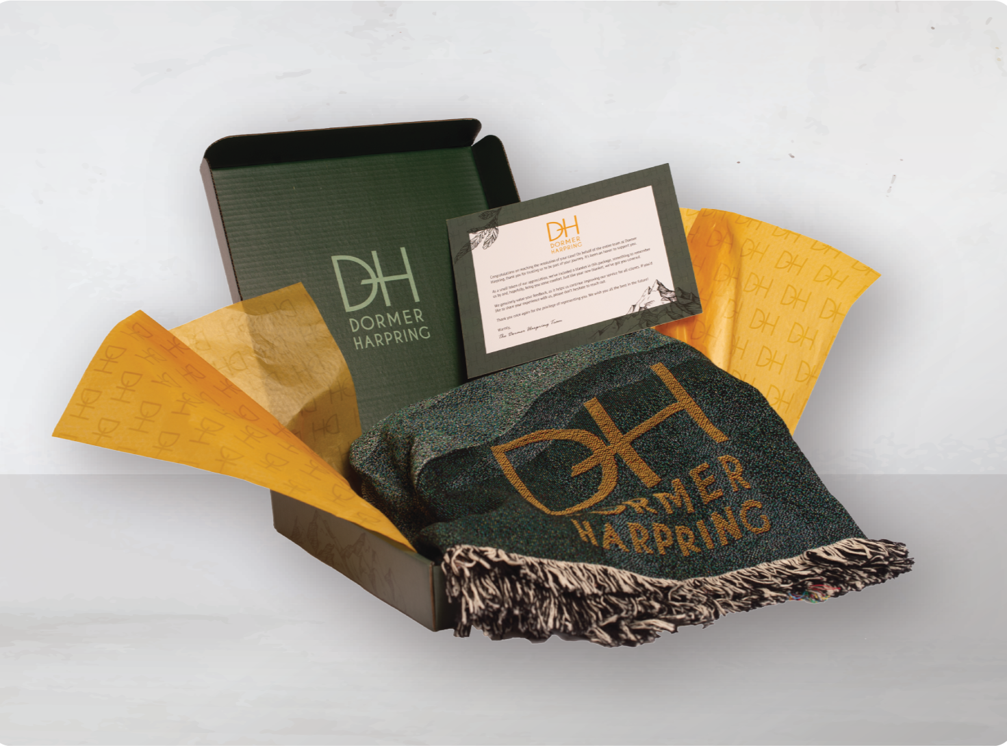 Open gift box with green interior, containing yellow tissue paper with 'DH' logo, a piece of fabric with 'DH Dormer Harring' logo, and a card congratulating on a new home.