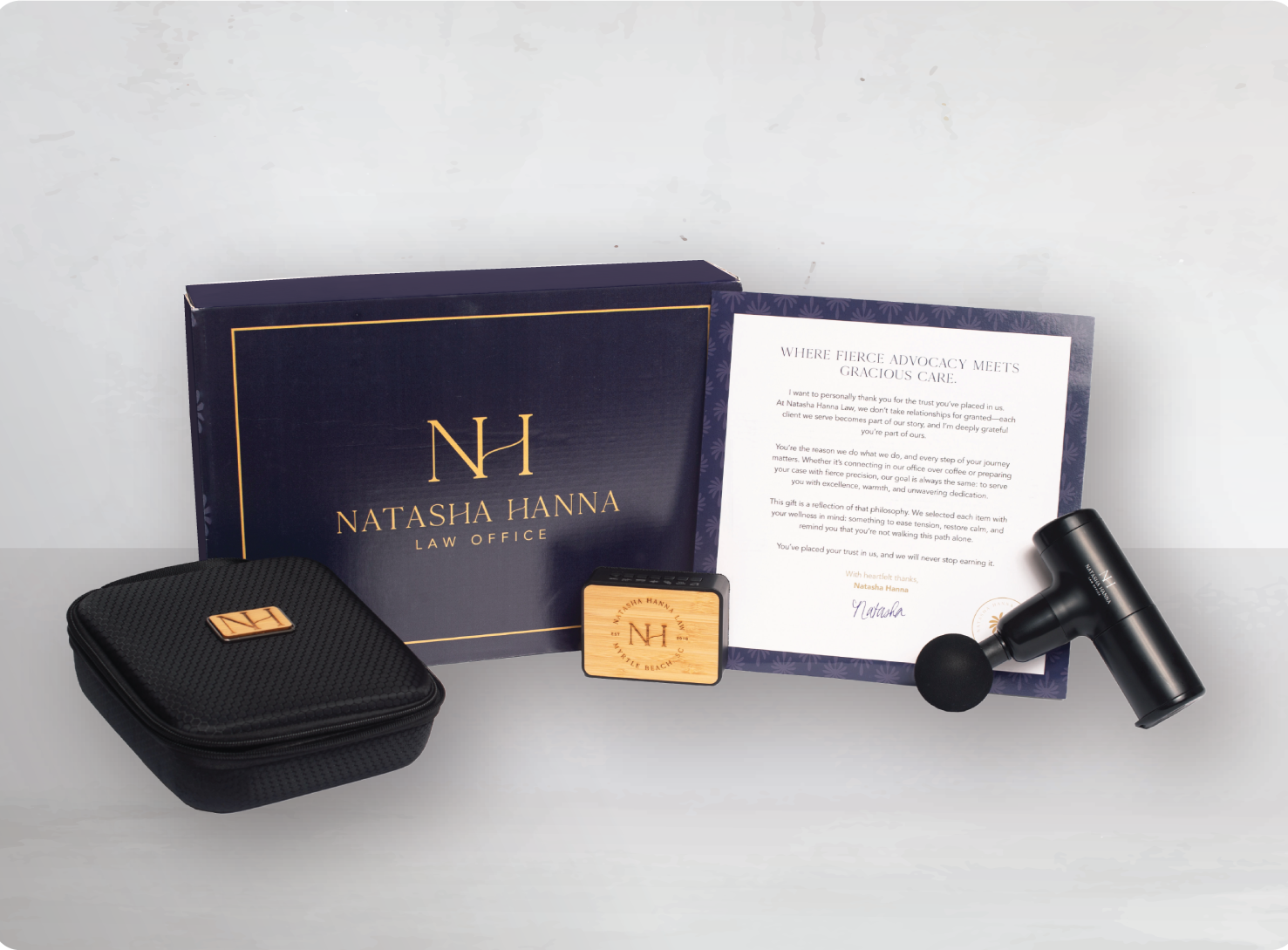Collection of promotional items for Natasha Hanna Law Office, including a navy blue box, a letter, a black massage gun, a wooden stamp, and a black zippered case with the law office's logo, all arranged on a white surface.