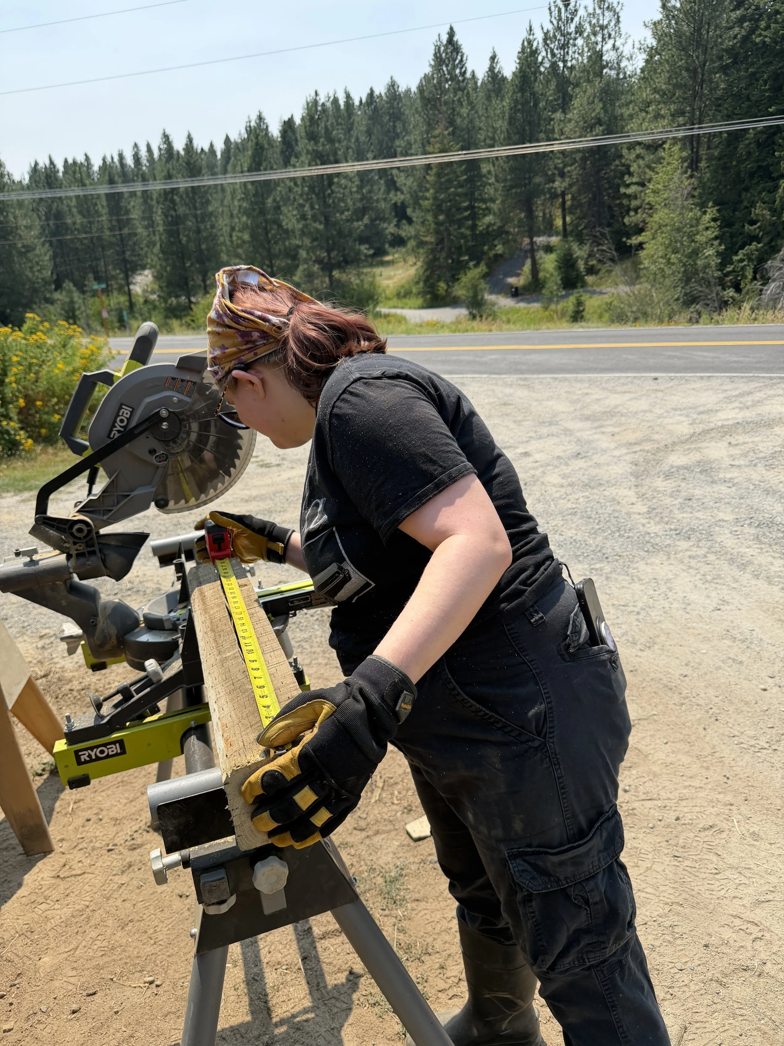 A woman with a patterned bandana and black clothing cutting wood with a large Ryobi chop saw outdoors on a gravel and dirt surface, with green trees and a road in the background.