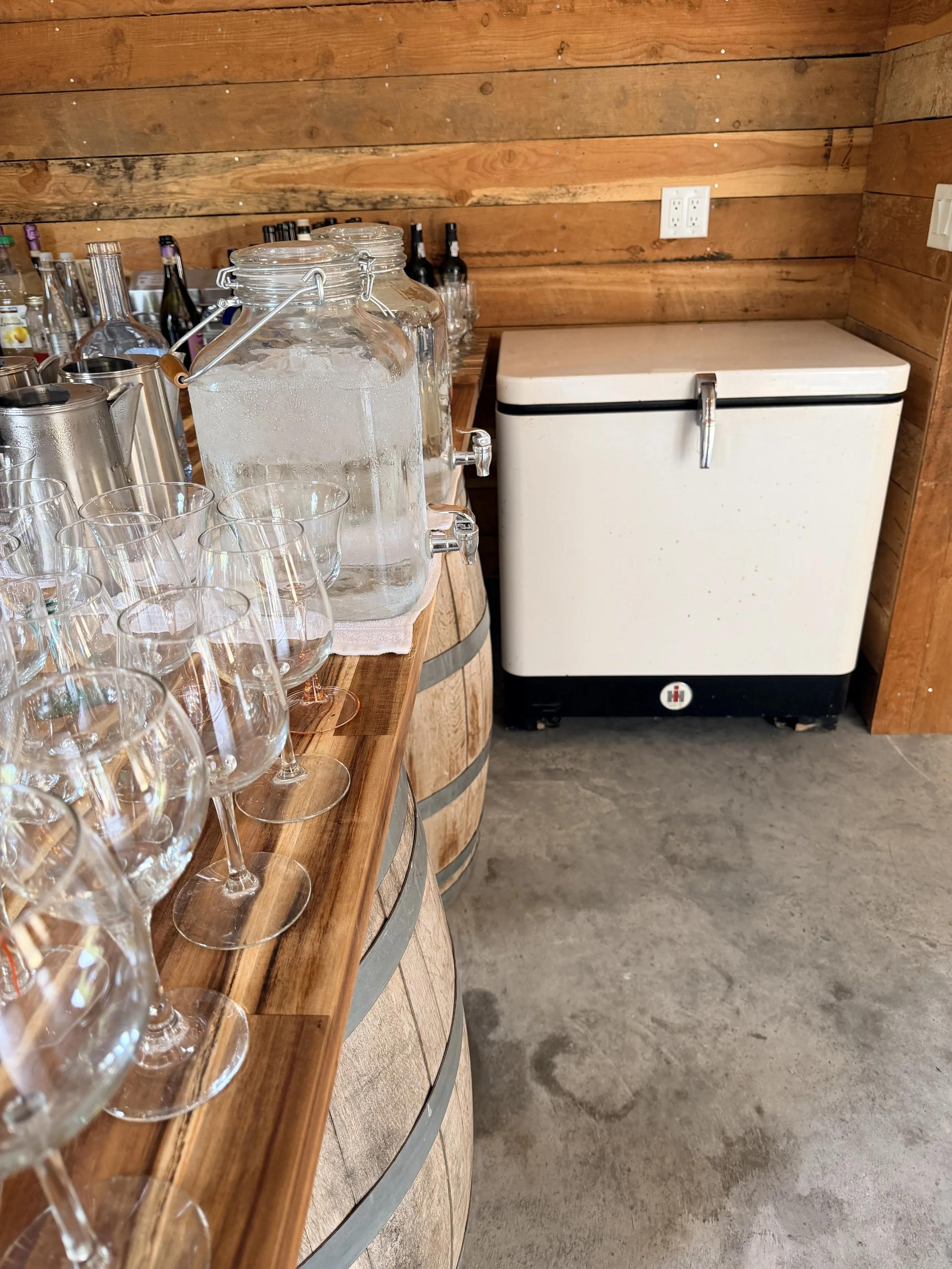 A wooden bar counter with several glasses and large glass dispensers filled with water, behind which are bottles of various alcoholic beverages. To the right, a white freezer with a latch, set against a wooden wall with an electrical outlet.