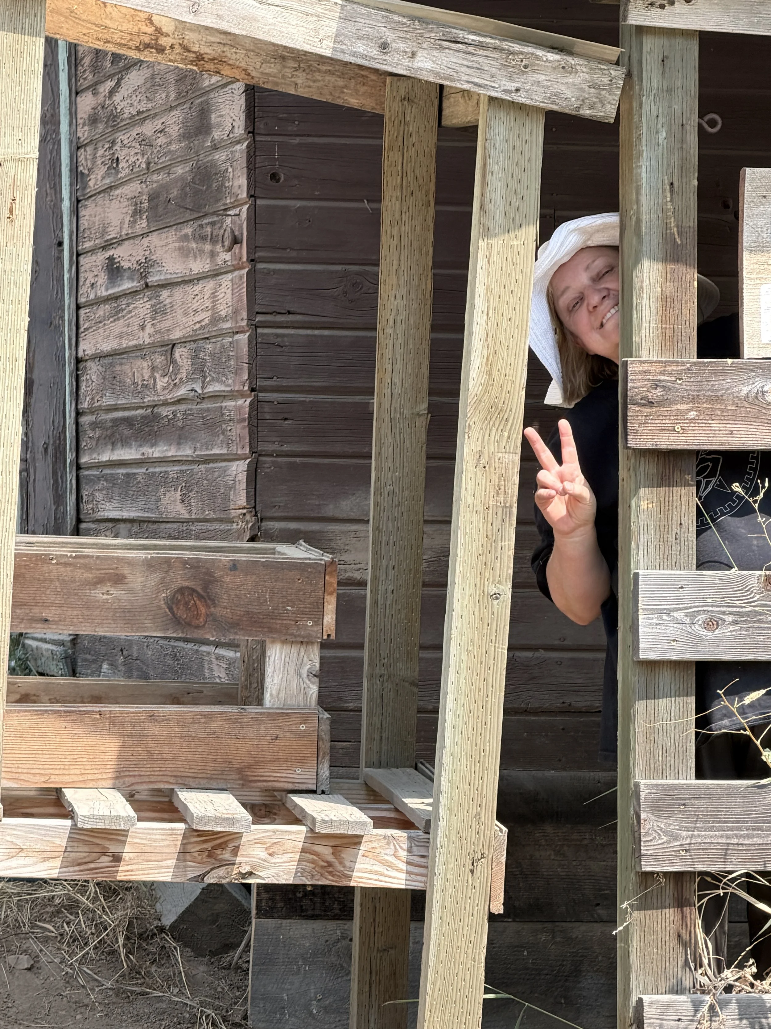 A woman wearing a white sun hat and black shirt is peeking through a gap in a wooden structure and flashing a peace sign with her fingers.