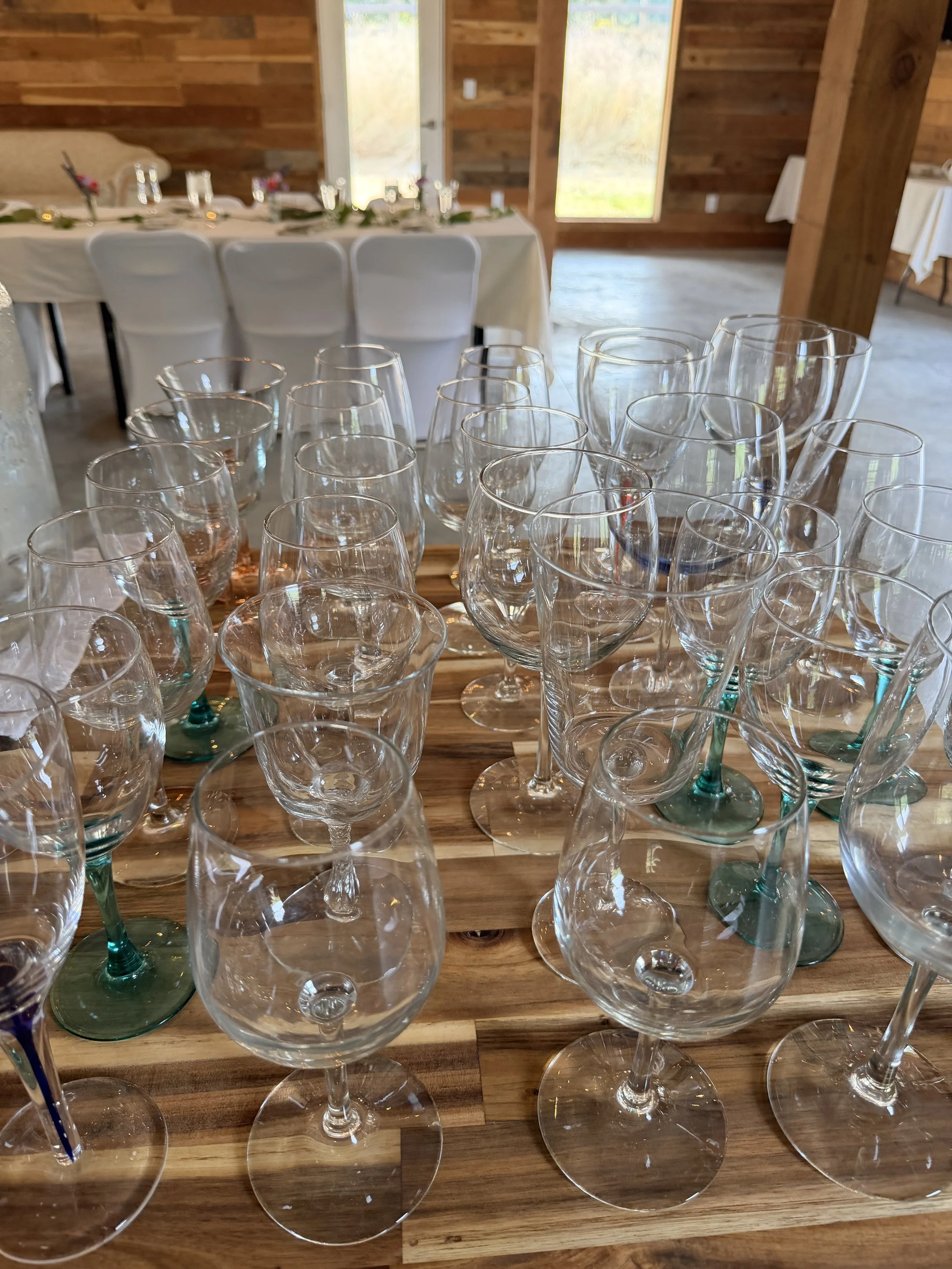 A collection of empty wine glasses on a wooden table in a rustic wooden room with a long decorated table set for a meal in the background.